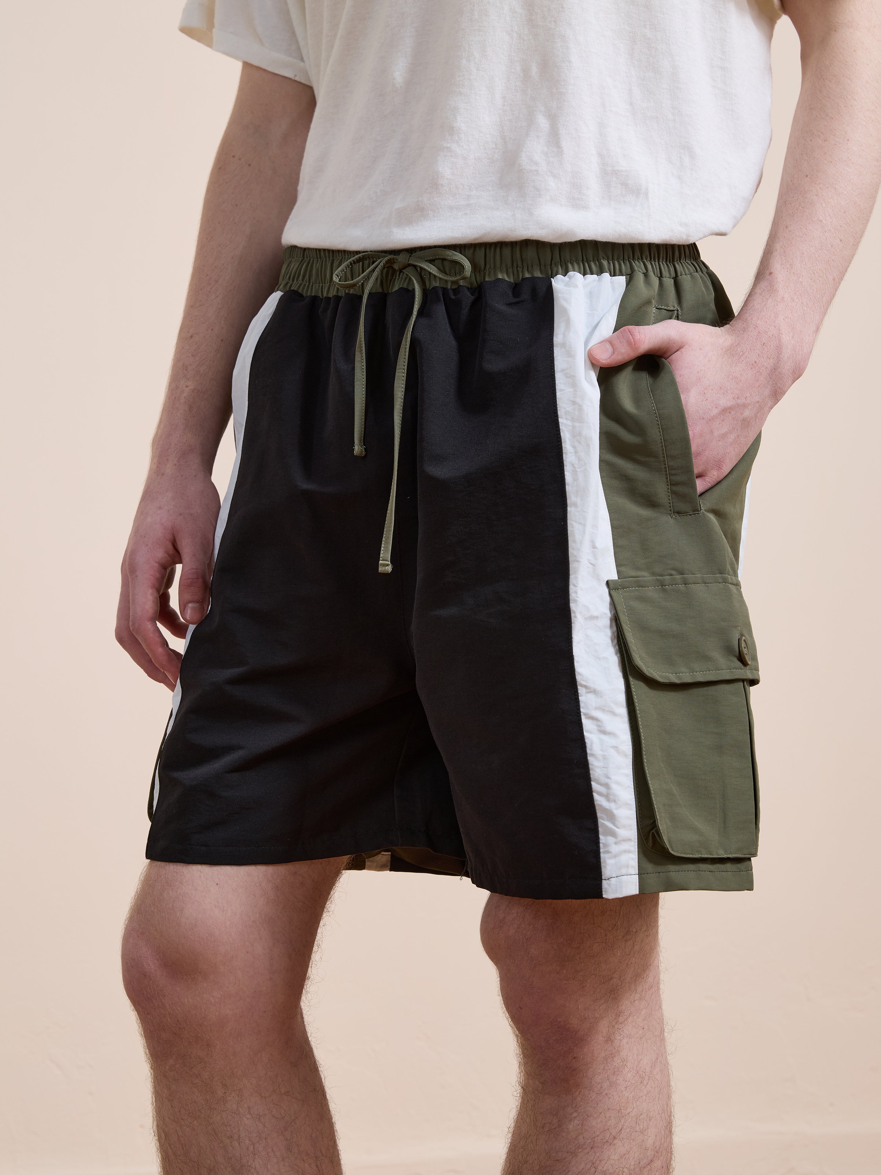 A person wears Found Sporting Club Track Shorts—vintage athletic, color-blocked black, white, and olive green shorts with a drawstring waist and cargo pocket—and a white t-shirt.