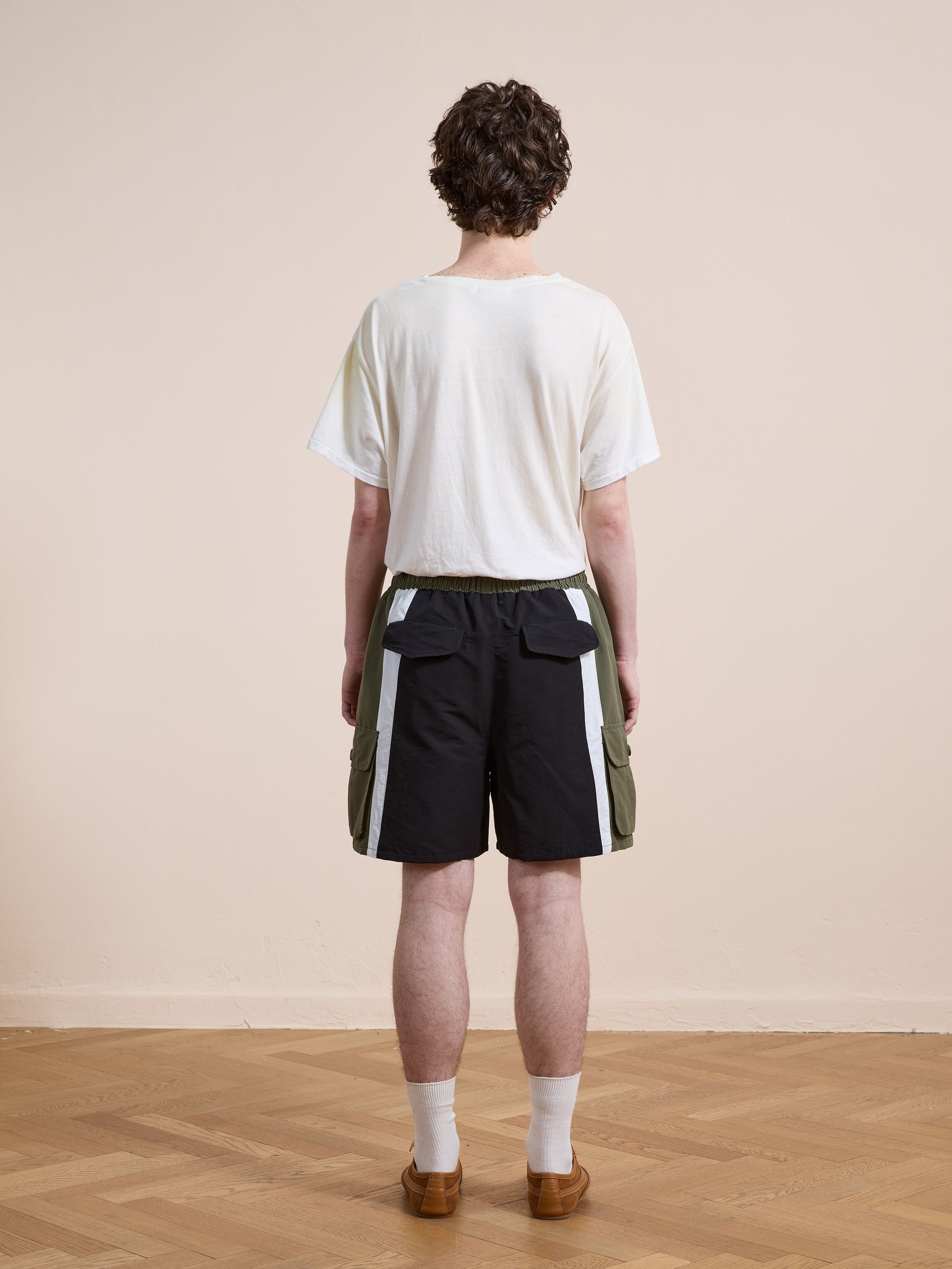 A person stands facing away, wearing a white t-shirt and Found Sporting Club Track Shorts with pocket details, paired with white socks and brown shoes on a wooden floor.