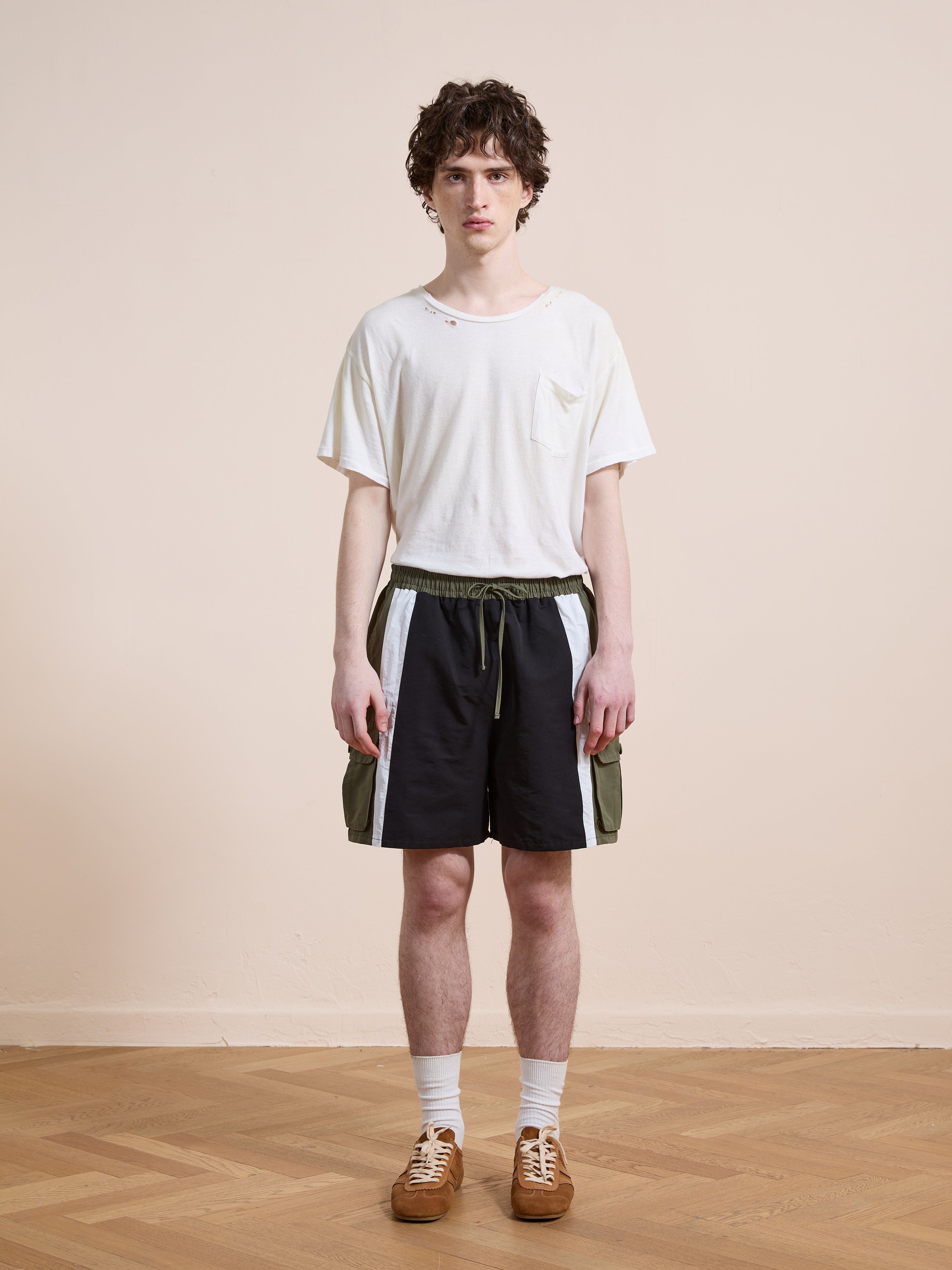 A young man with curly hair stands indoors, facing forward in a white t-shirt, Found Sporting Club Track Shorts, white socks, and brown sneakers. He wears a neutral expression.