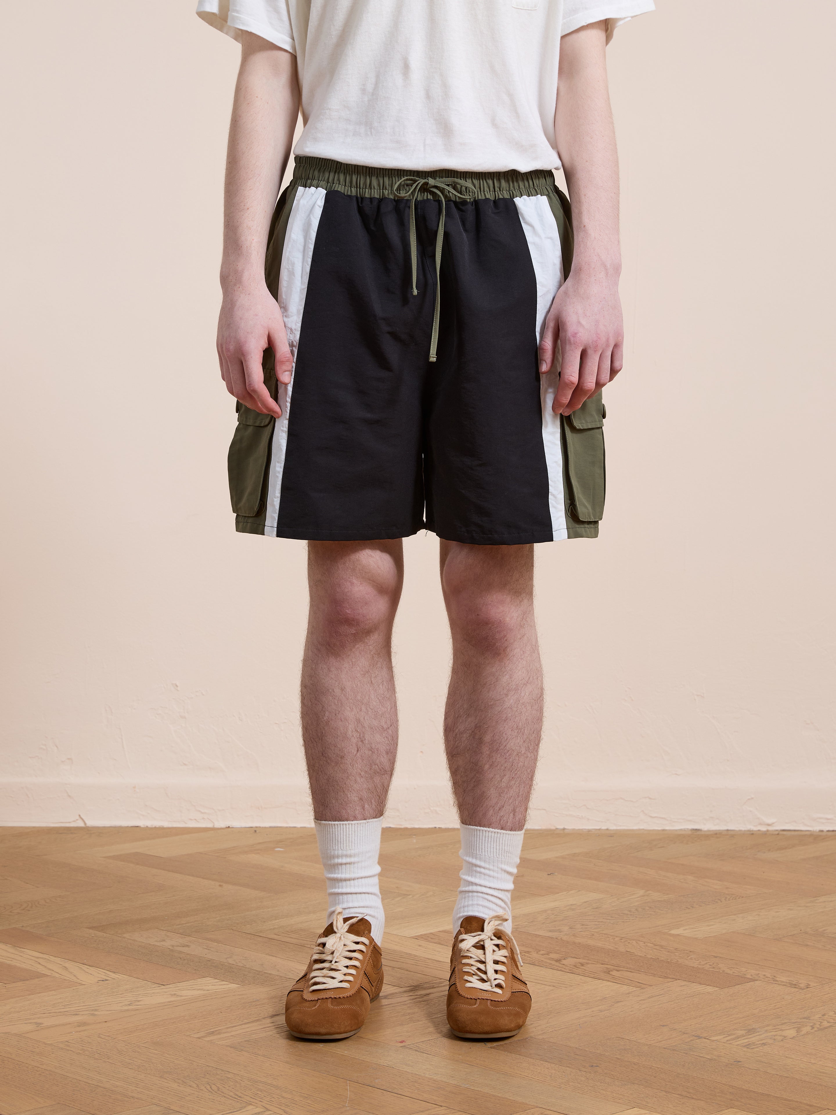 A person wearing a white t-shirt, Found Sporting Club Track Shorts with green pockets, white socks, and brown sneakers stands on a wooden floor against a plain wall.