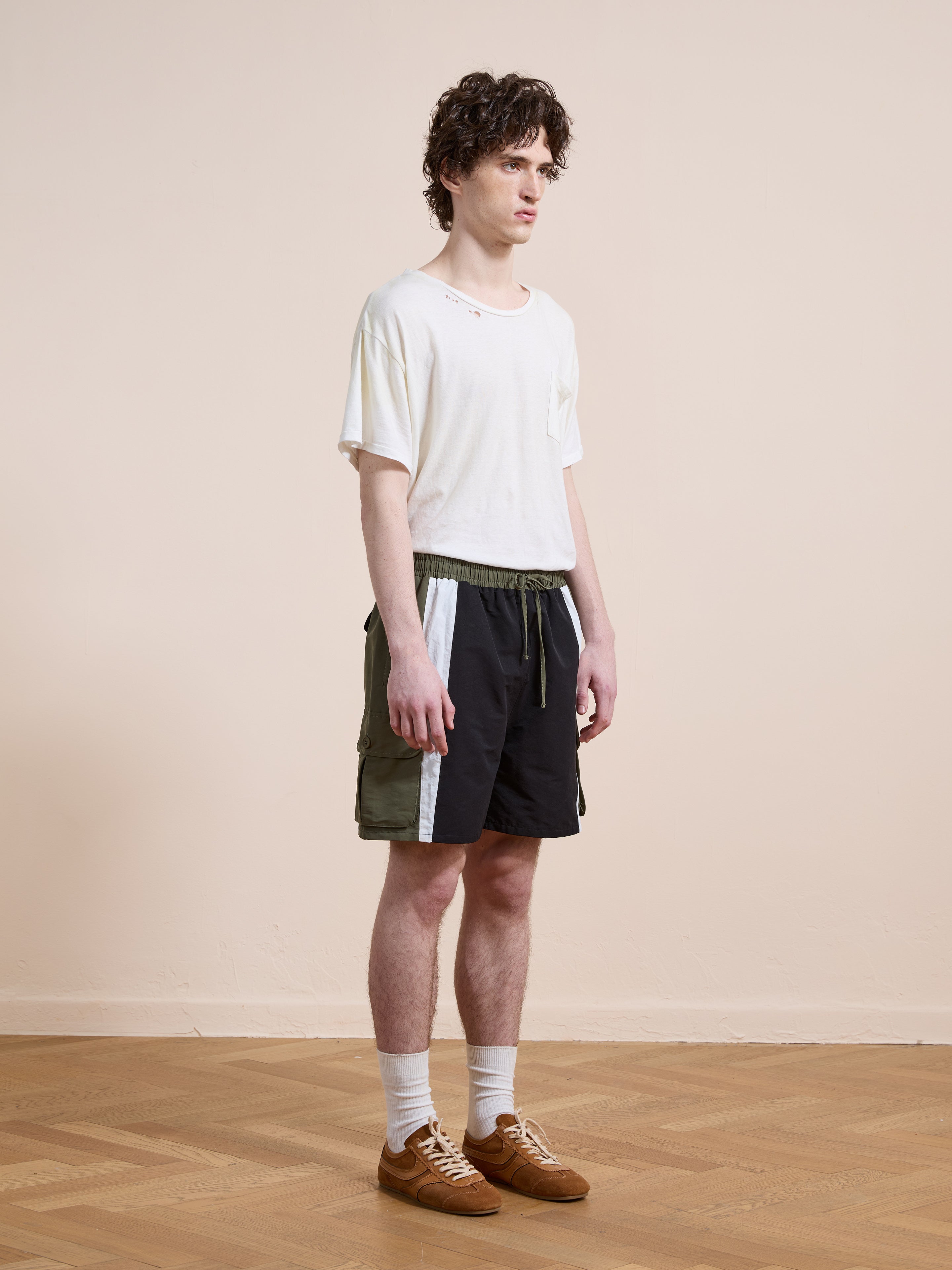 A young man stands on a wooden floor, wearing a white t-shirt, Found Sporting Club Track Shorts with white stripes, white socks, and brown sneakers for a classic unisex look.