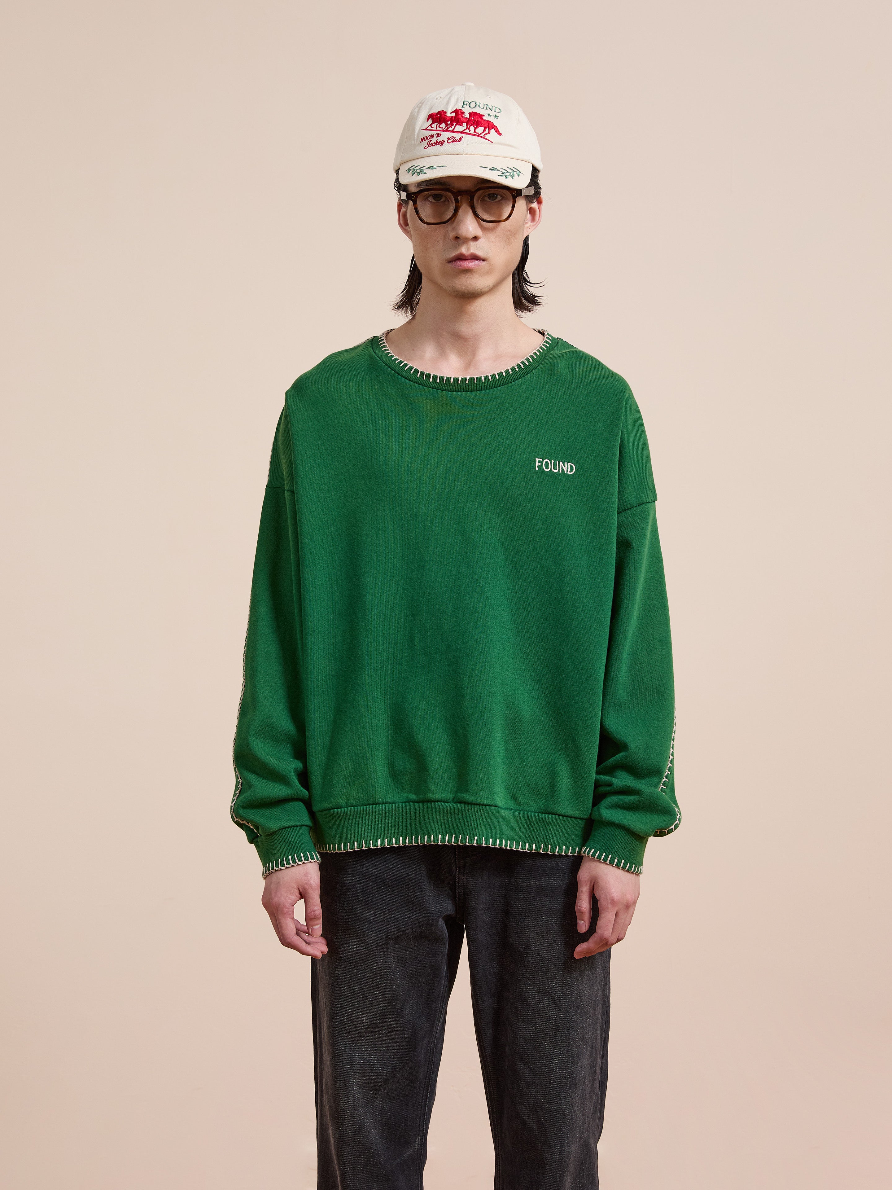 A person in a green FOUND Stitched Border Distressed Crewneck made from French terry cotton, with dark jeans, glasses, and a white cap stands against a plain beige background.