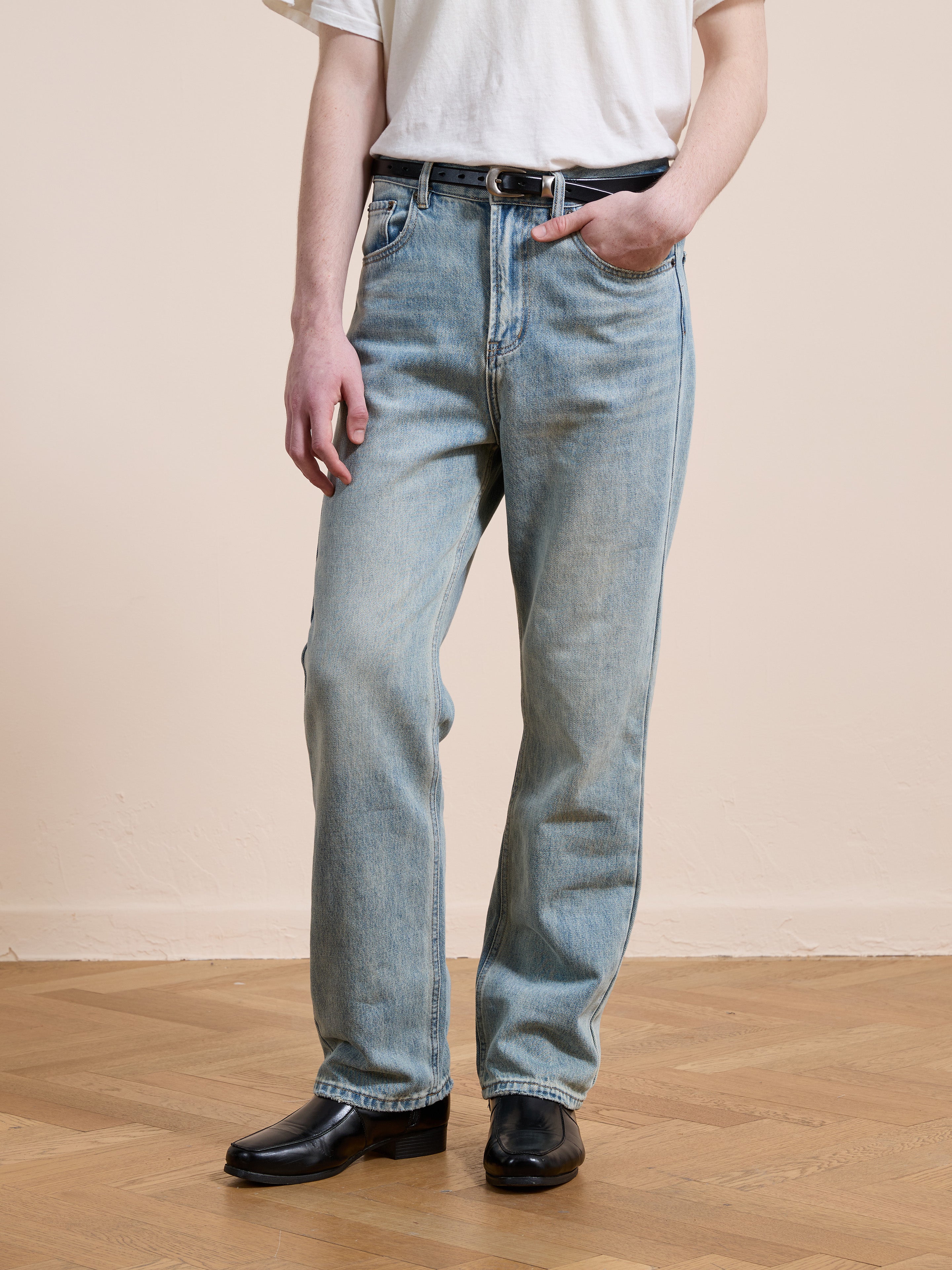 A person in a white t-shirt, black shoes, and Found Straight Leg Jeans stands on a wooden floor with one hand in their pocket.
