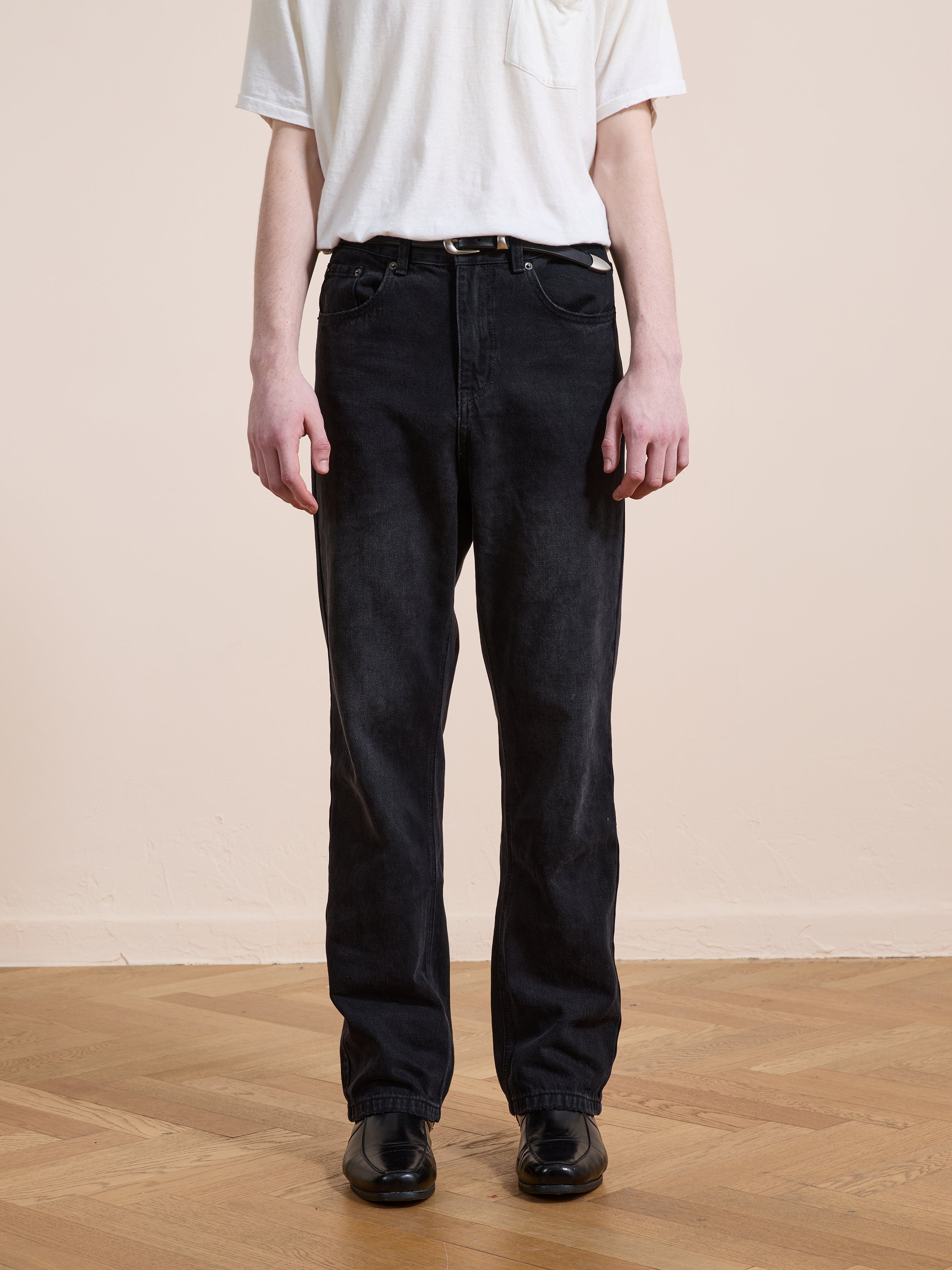 Someone wearing a white T-shirt tucked into Found Straight Leg Jeans in black cotton denim, styled with a black belt and shoes, stands on a wooden floor against a plain light-colored wall.