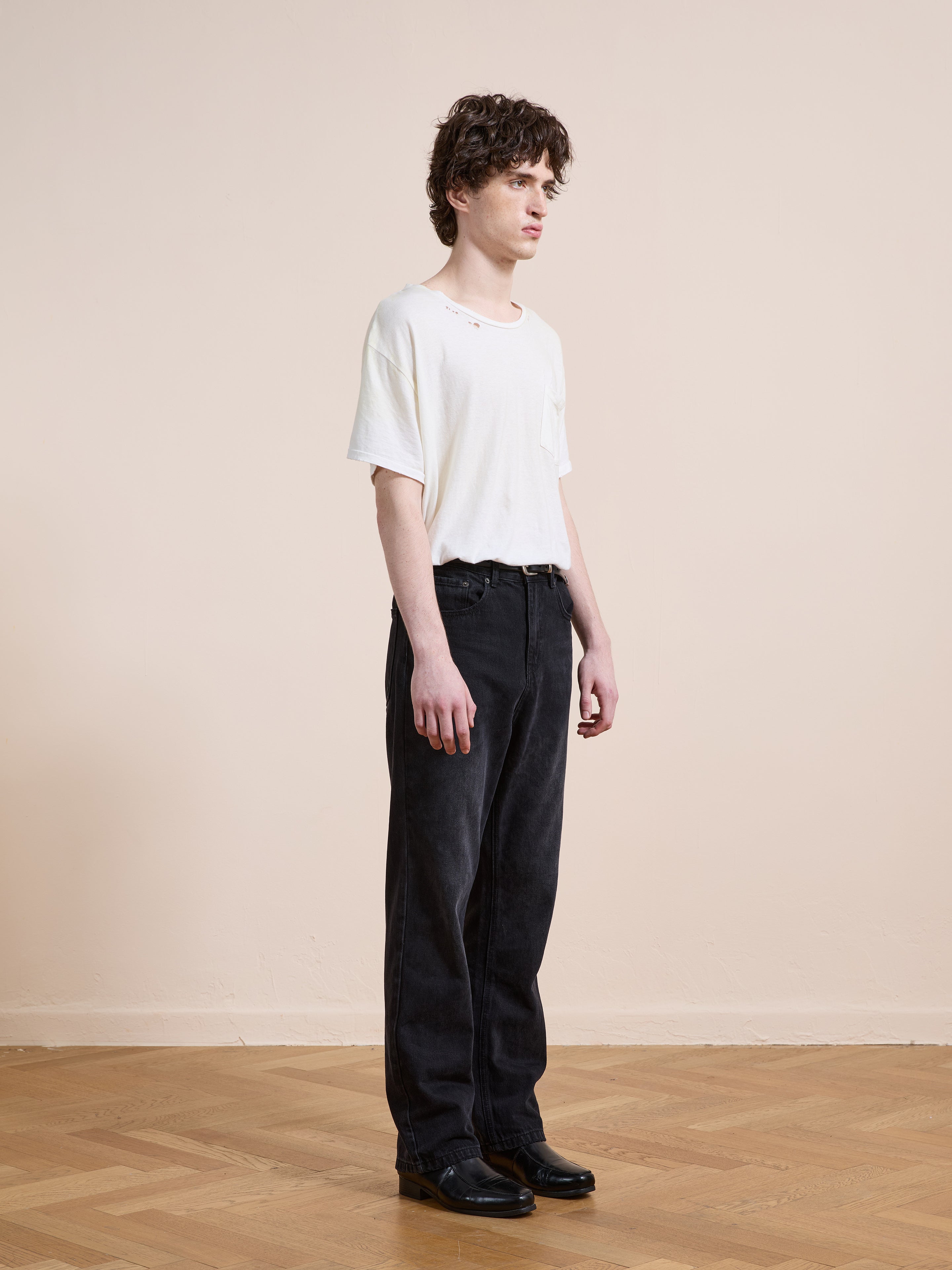 A person stands indoors against a plain beige wall, wearing a white t-shirt, black Found Straight Leg Jeans made from cotton denim, and black shoes, looking to the side with a neutral expression.