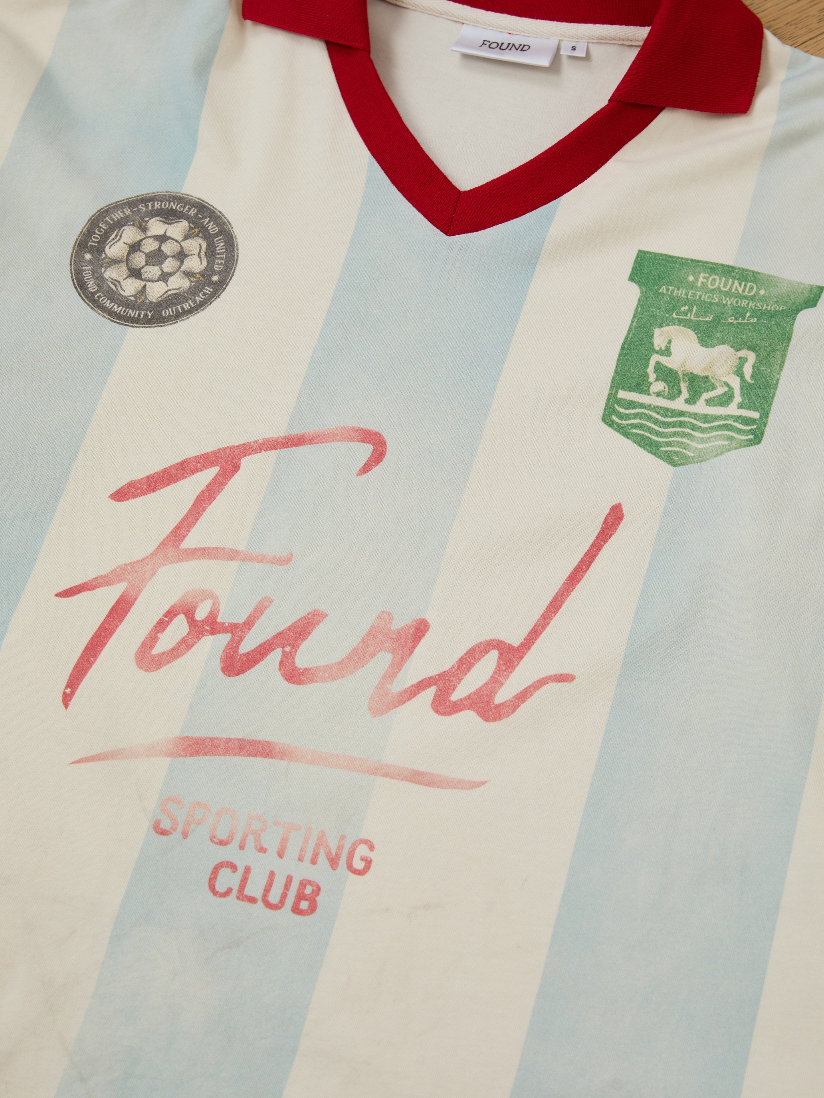 The Striped Sport LS Jersey by FOUND is a vintage-inspired sportswear piece featuring light blue stripes and a red collar. With "Found Sporting Club" on the front, it's ideal for football enthusiasts who appreciate classic style.