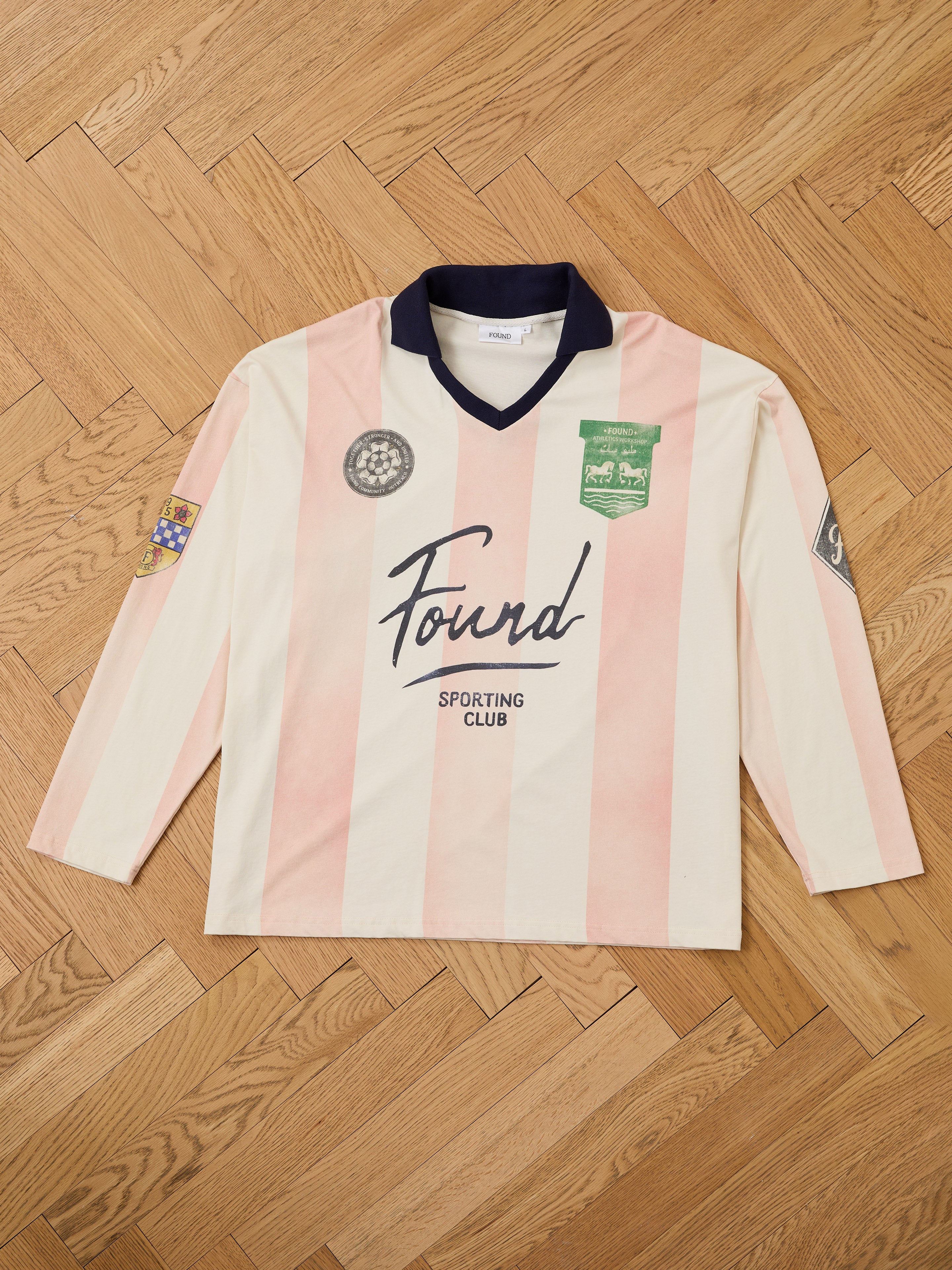 The FOUND Striped Sport LS Jersey is a unisex, relaxed-fit long-sleeve jersey featuring pink and cream vertical stripes, a navy collar, and multiple embroidered patches, shown laid flat on a wooden herringbone floor.