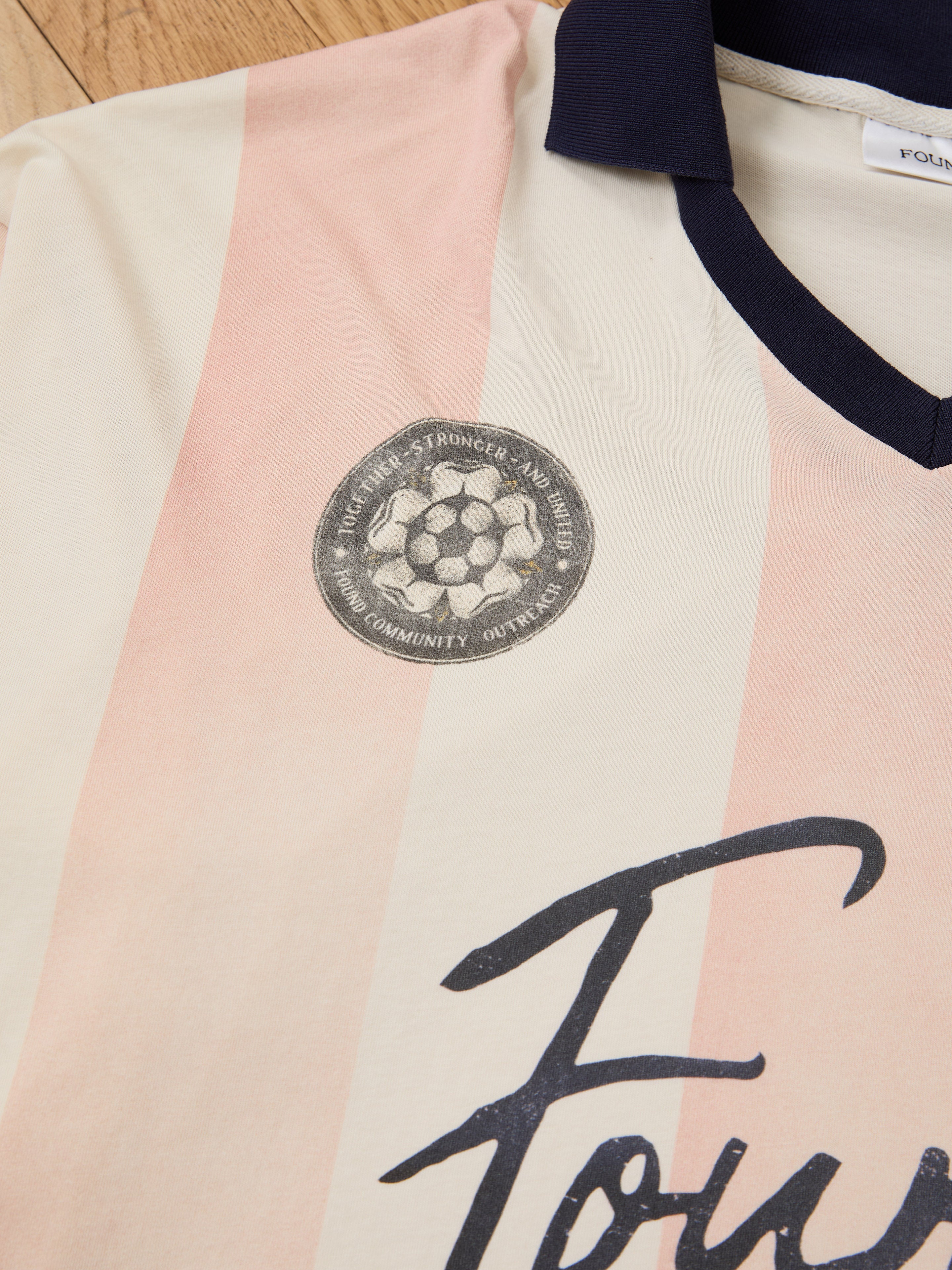 A close-up of the FOUND Striped Sport LS Jersey, featuring cream and pink stripes, a dark collar, round soccer emblem, cursive “Four” on the chest, and a unisex relaxed fit.