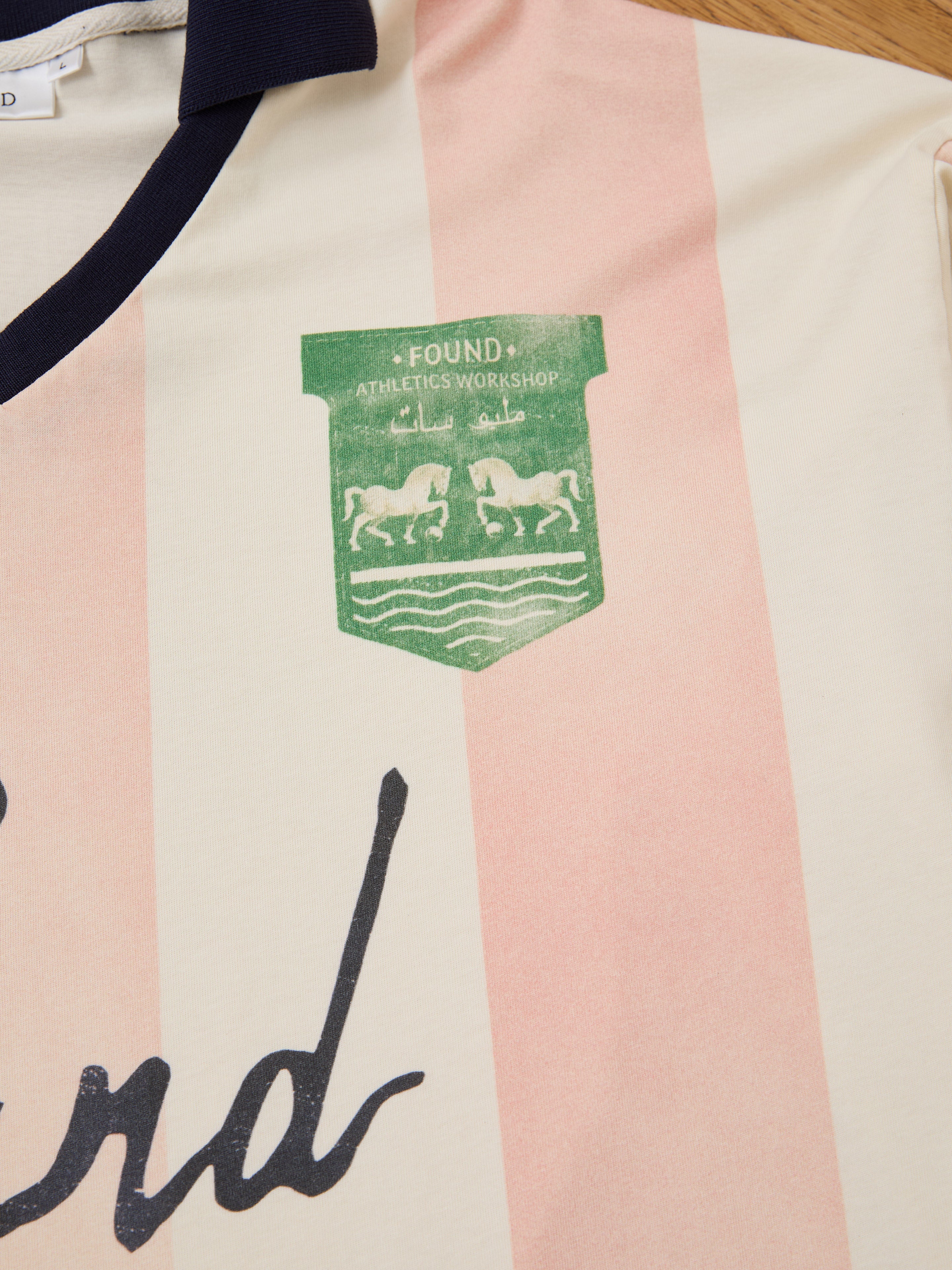 Close-up of the FOUND Striped Sport LS Jersey, a unisex relaxed-fit shirt with pink and off-white stripes, featuring a green crest with two horses, waves, and "FOUND ATHLETICS WORKSHOP" in English and Arabic.