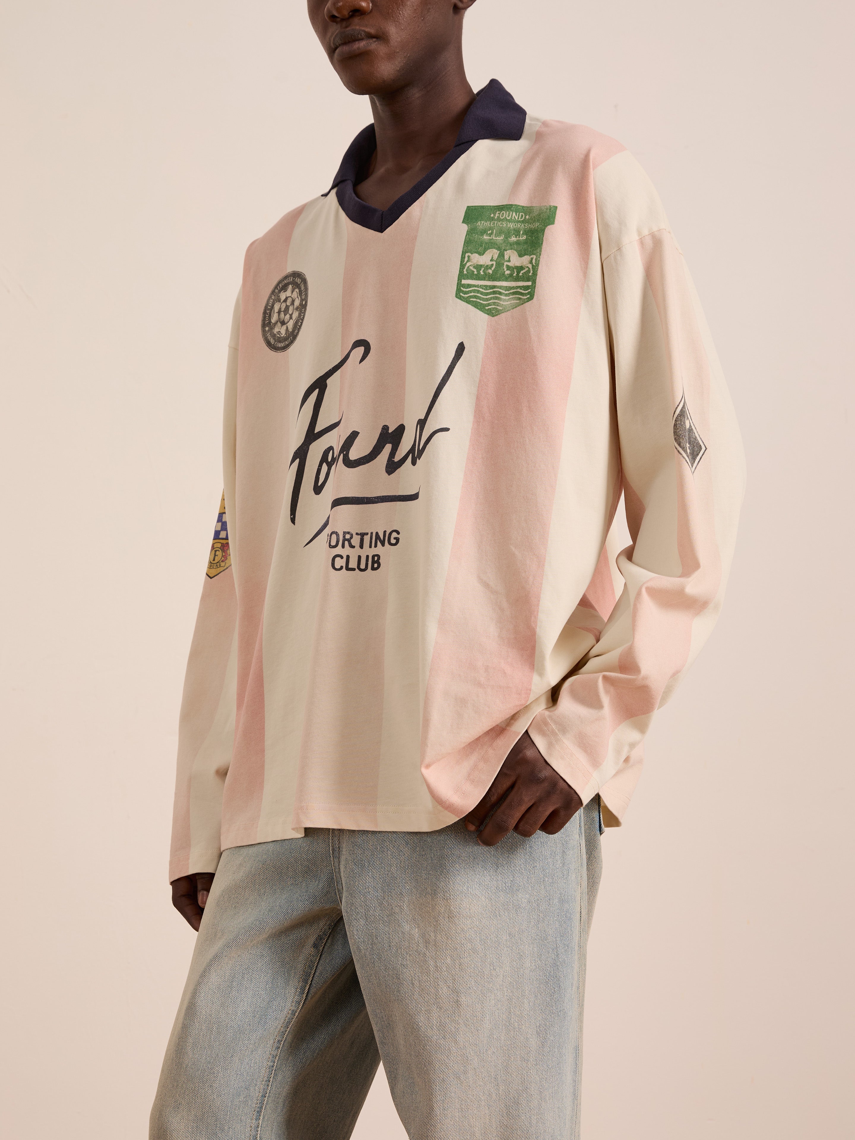 Someone wearing a loose, long-sleeve, pink and cream FOUND Striped Sport LS Jersey with multiple patches and logos, styled with light blue jeans for a relaxed unisex fit.