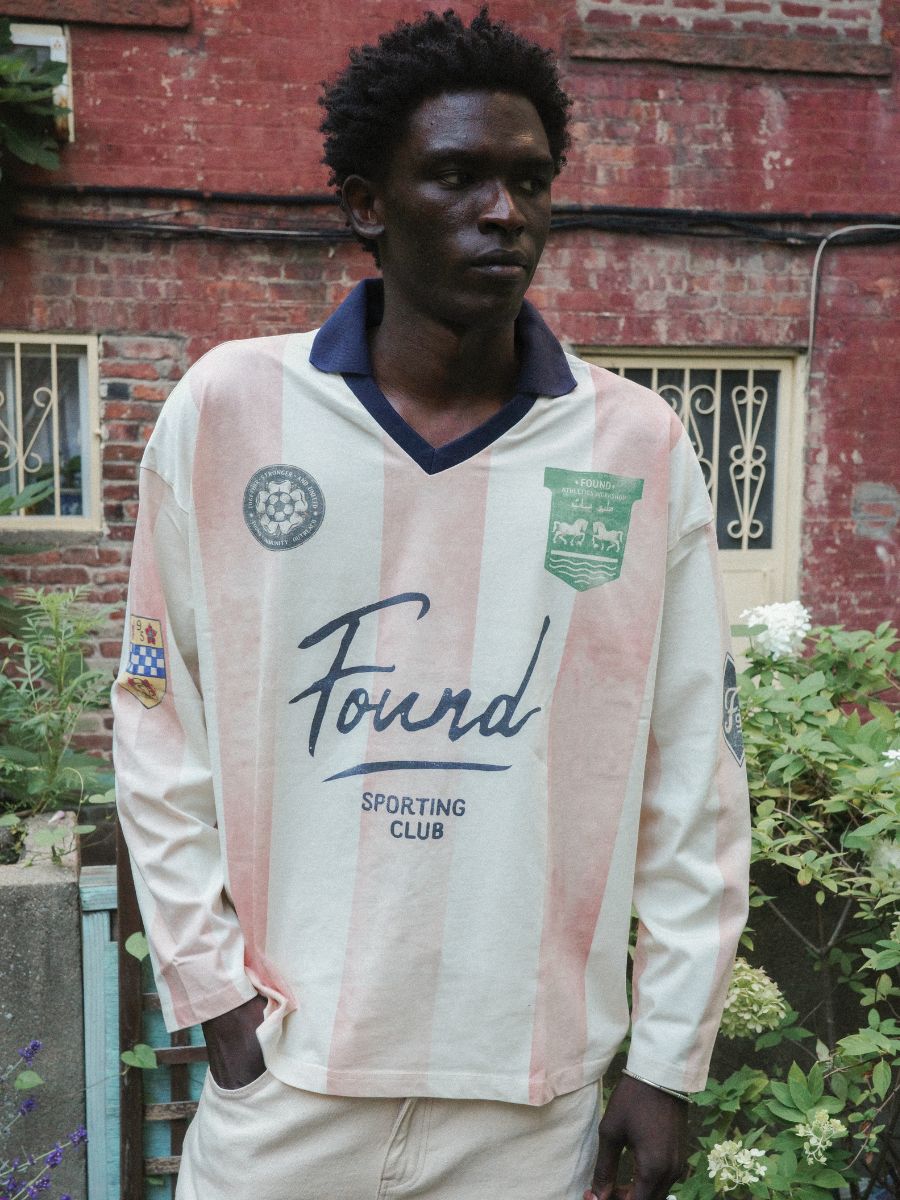 A man stands outside in the FOUND Striped Sport LS Jersey, featuring cream and pink stripes, a unisex relaxed fit, logo, and patches—with a brick building and garden in the background.