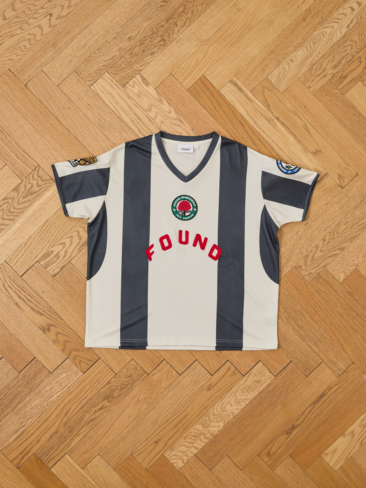 The Striped Stateman Jersey by FOUND features wide dark stripes, red “FOUND” text, and sleeve patches—shown laid flat on a wooden floor.