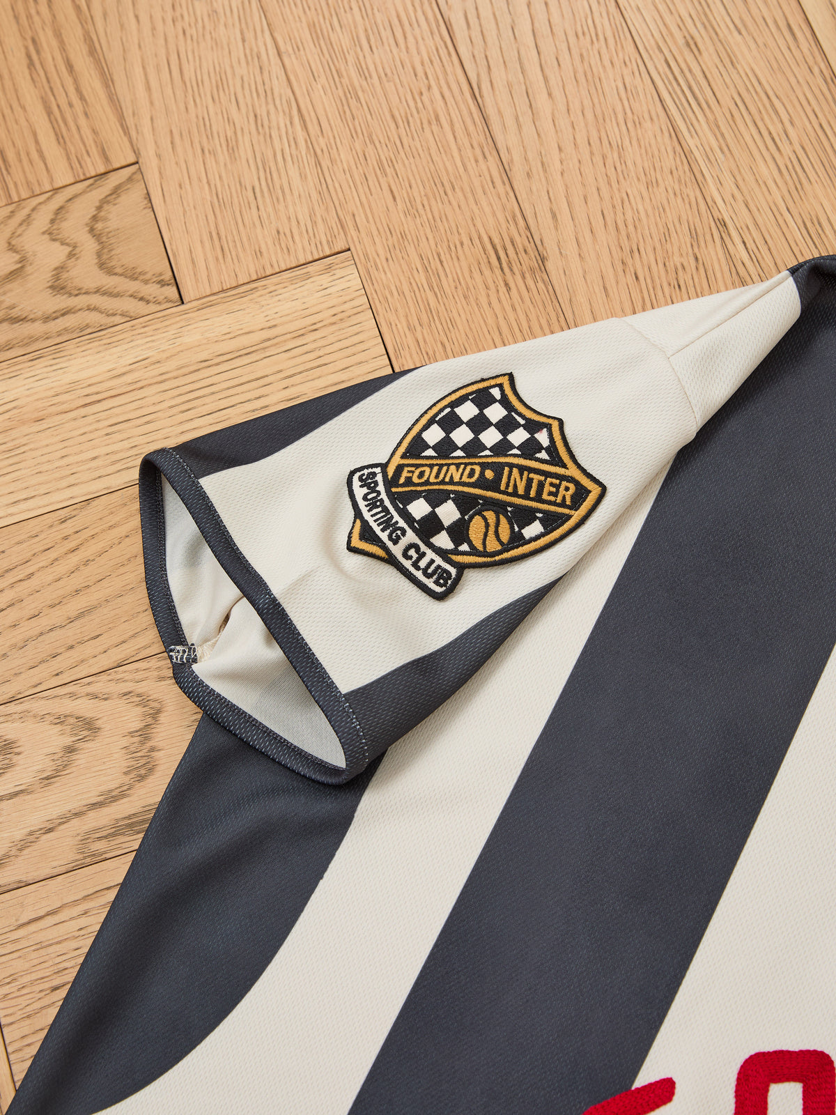 Close-up of the Striped Stateman Jersey by FOUND, a black and white vintage-style football shirt with a checkered shield patch, on a wooden floor.