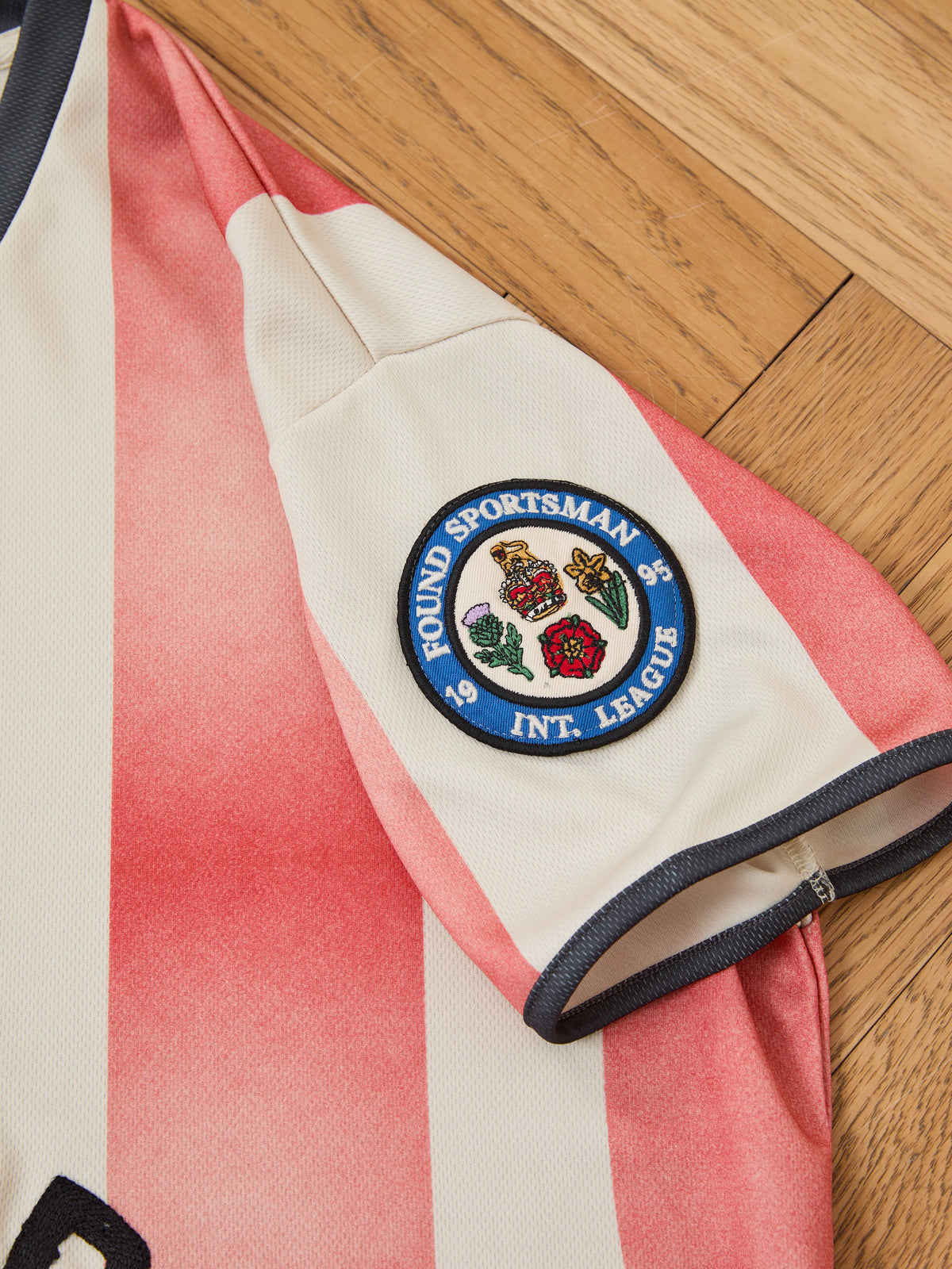 The FOUND Striped Stateman Jersey—a short-sleeved shirt with red and white stripes inspired by vintage football jerseys—features a round patch on the sleeve that reads “Pound Sportsman Int. League” with crests in the center, and is displayed on a wooden floor.