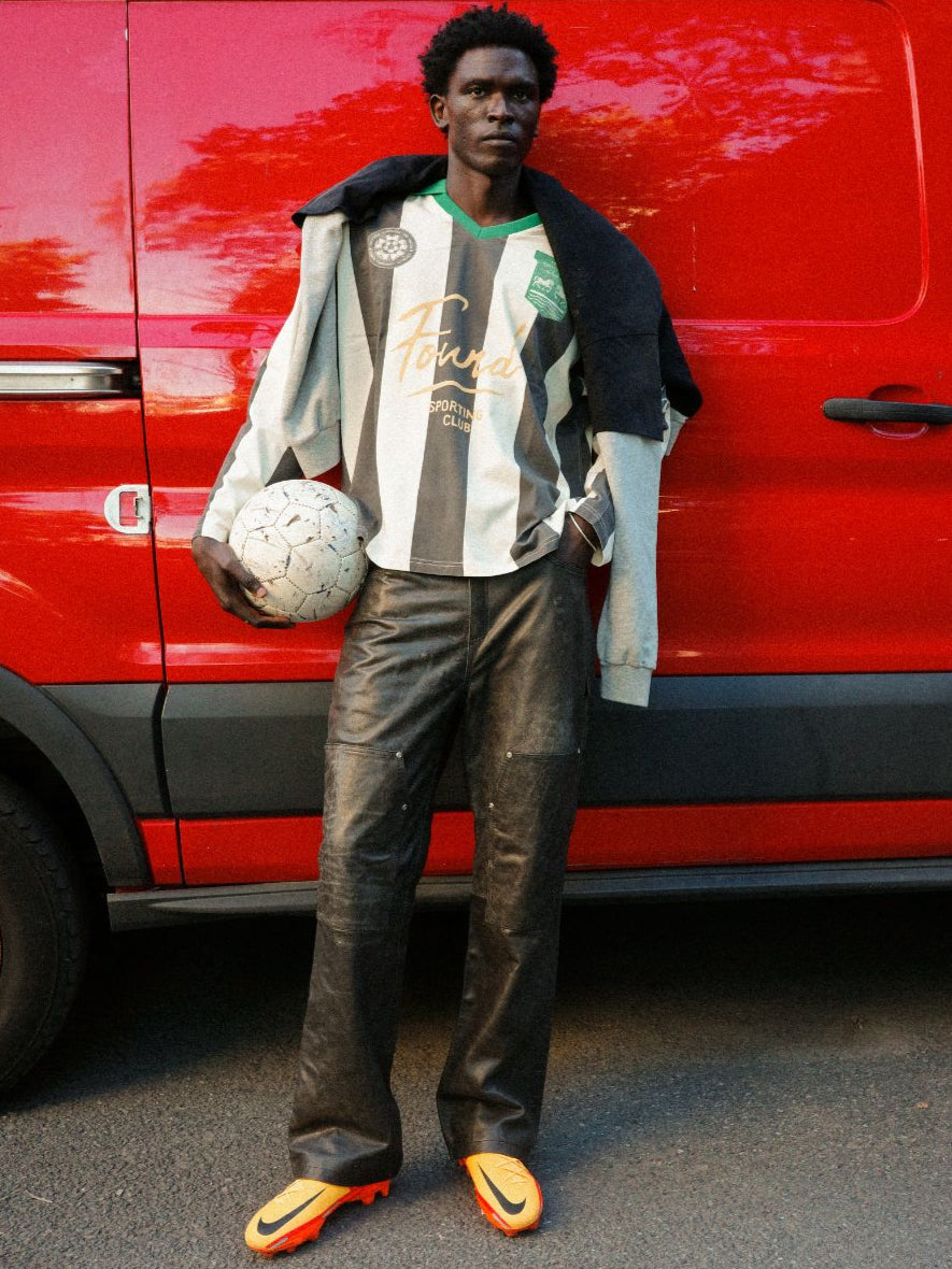 Wearing the FOUND Striped Sport LS Jersey, a person in black and white stripes with leather pants and orange cleats holds a soccer ball in front of a red van. The unisex relaxed fit creates a sporty-casual look.