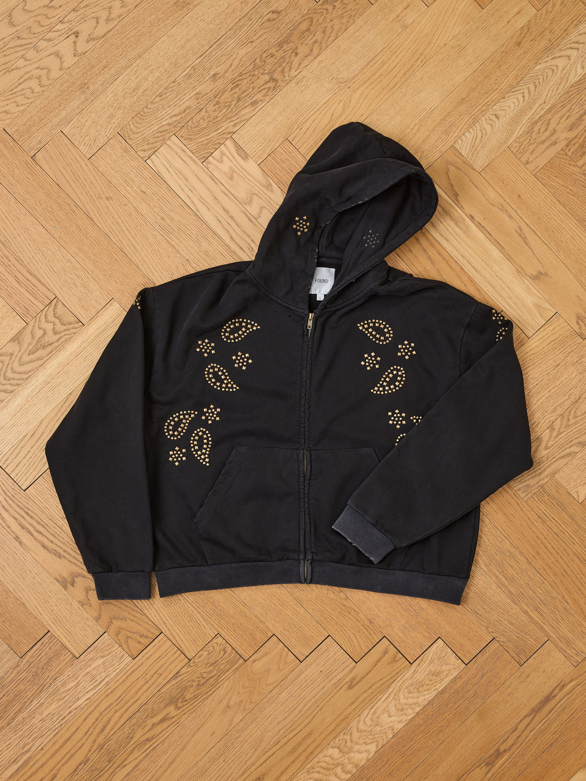 FOUND Studded Zip Up Hoodie with gold paisley designs, shown flat on a light brown parquet wooden floor.