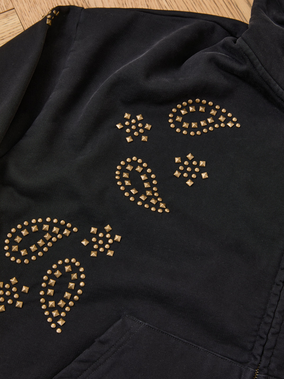 FOUND Studded Zip Up Hoodie—vintage black with gold square studs in bandana paisley patterns, laid on a wooden floor.