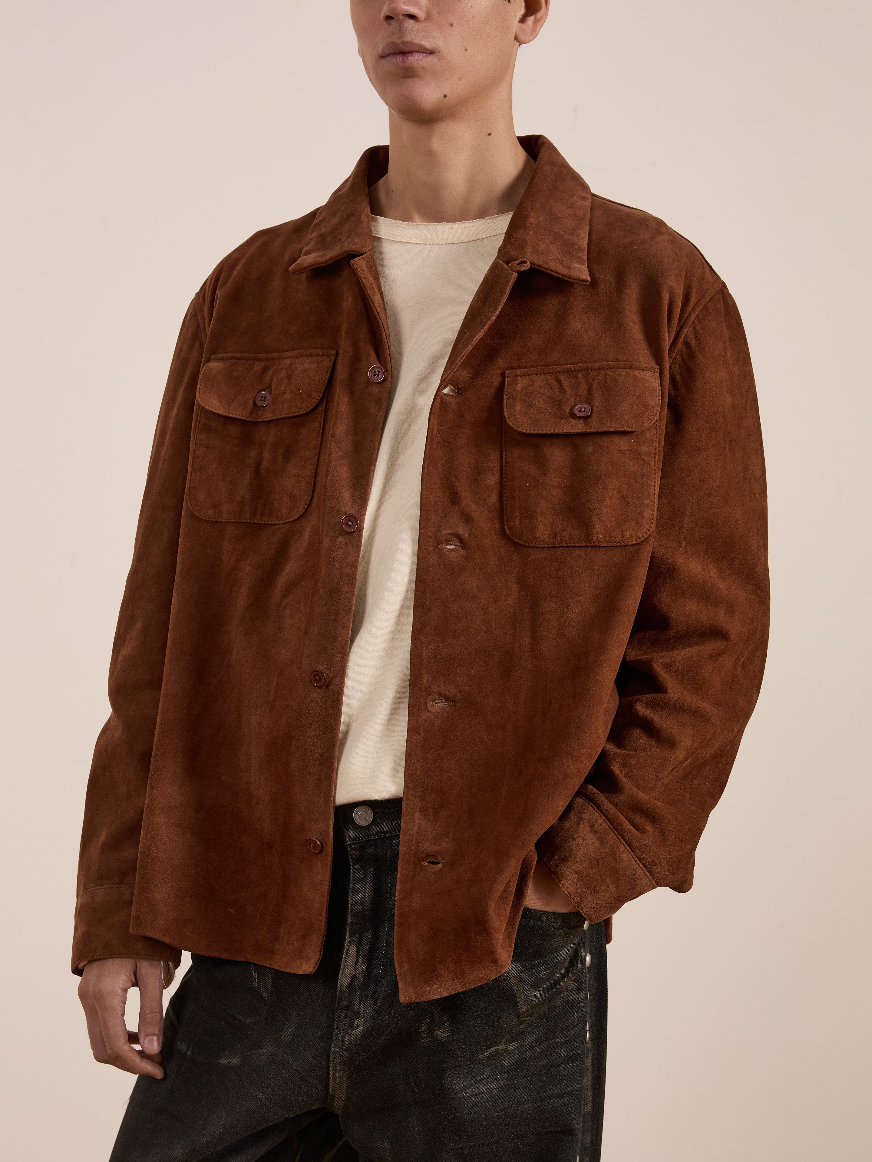A person wearing the FOUND Suede Leather Camp Shirt—a brown goat suede button-up with a unisex relaxed fit and two chest pockets—over a beige shirt and dark jeans, standing against a plain background.