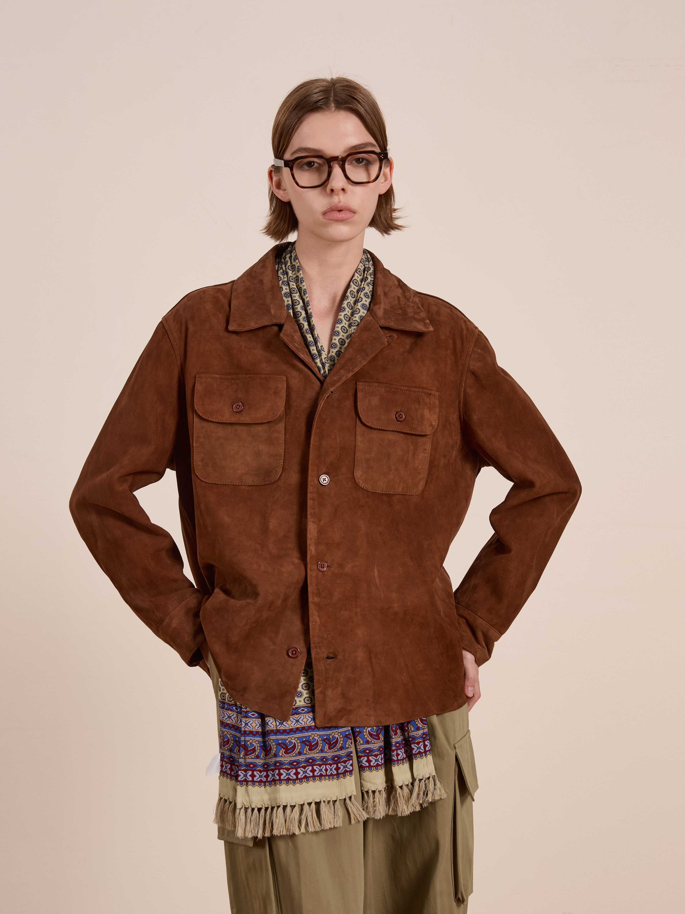 A person with long hair and glasses wears the FOUND Suede Leather Camp Shirt in a unisex relaxed fit, styled over a patterned scarf and olive green pants, standing against a plain light background.