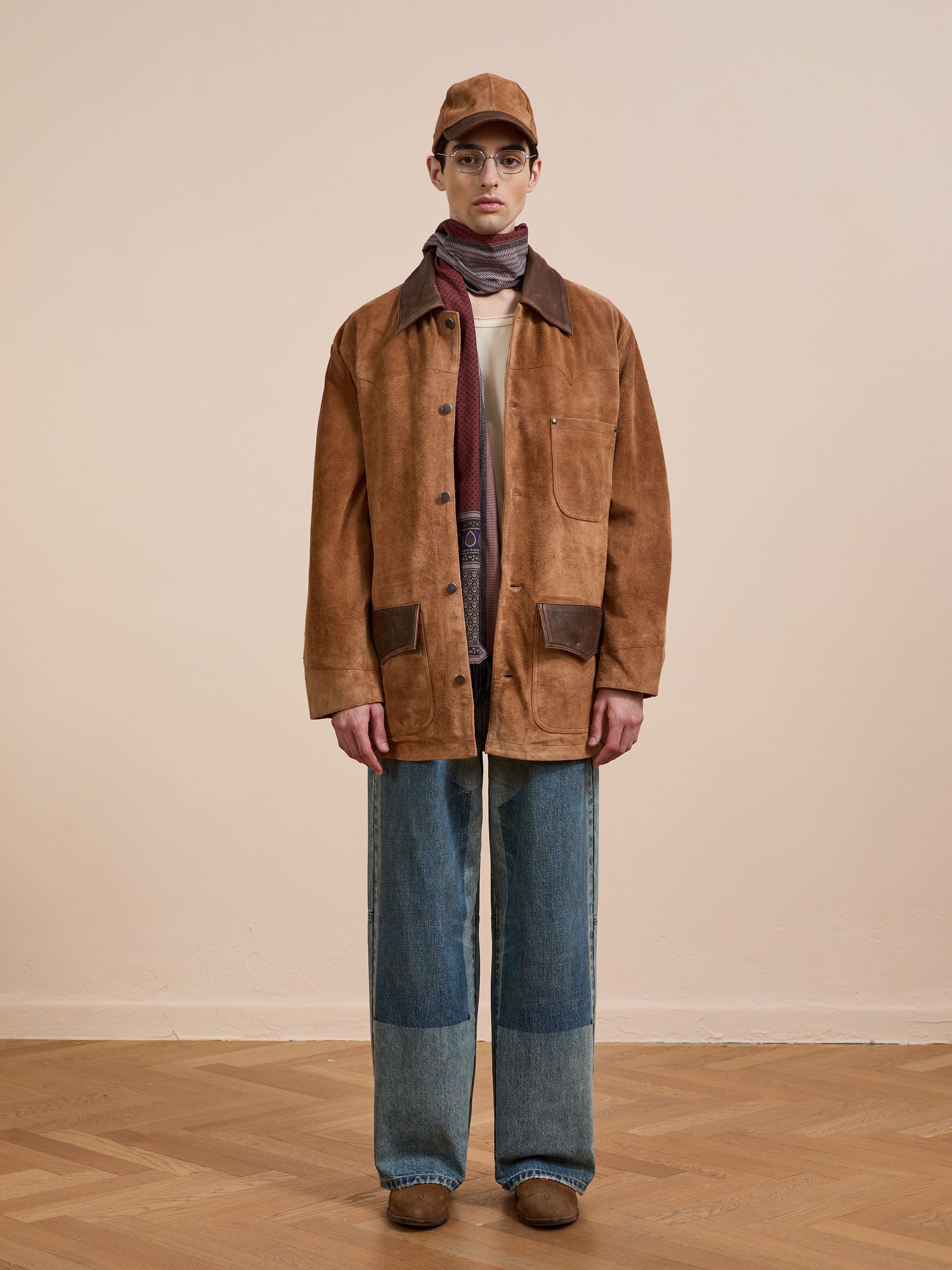 A person stands indoors by a plain wall wearing the FOUND Suede Contrast Chore Jacket, blue patchwork jeans, brown shoes, a beige cap, glasses, and a multi-colored scarf.
