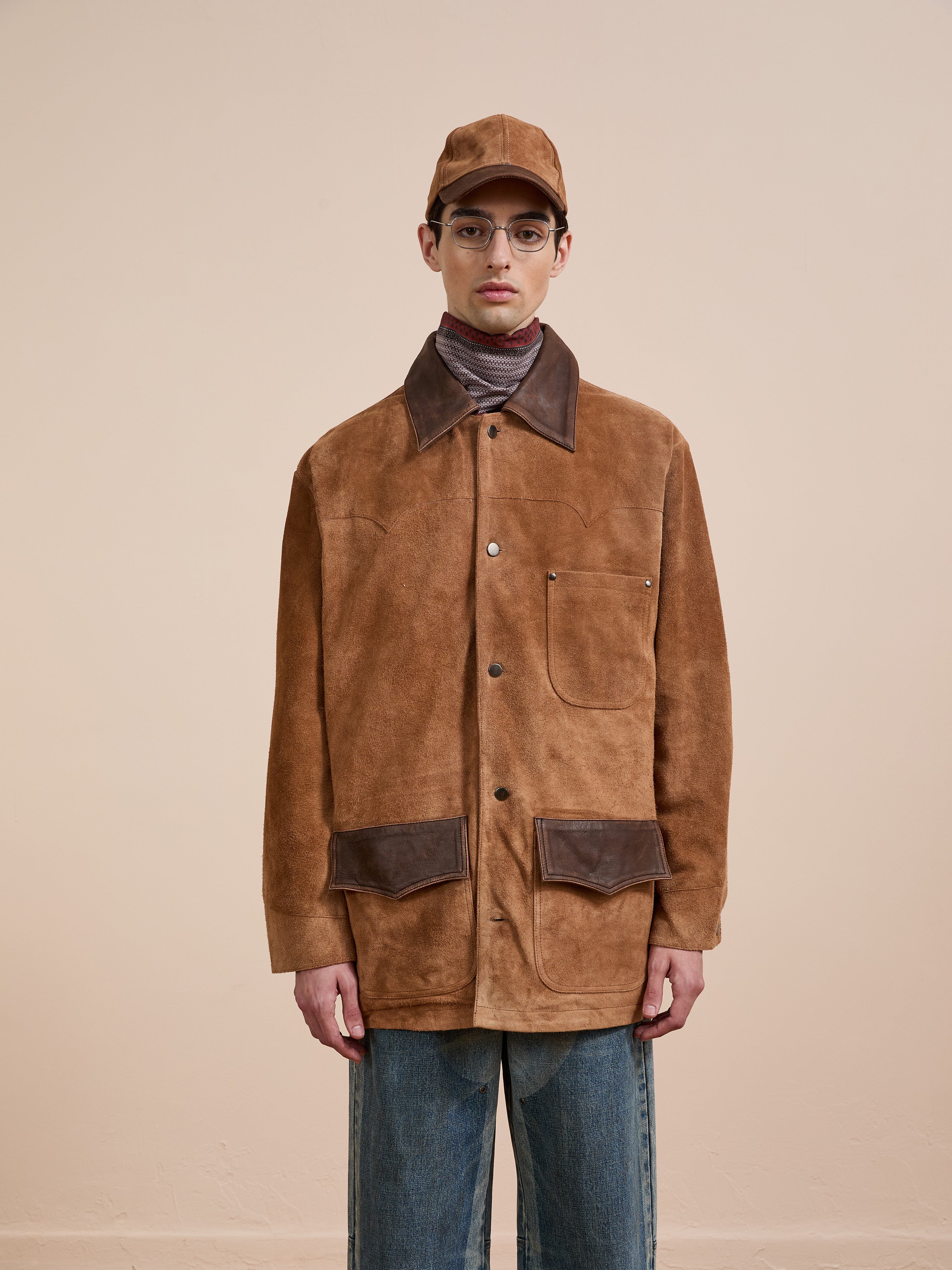 Wearing FOUND’s Suede Contrast Chore Jacket, blue jeans, a brown cap, glasses, and a turtleneck, a person stands against a plain beige background.