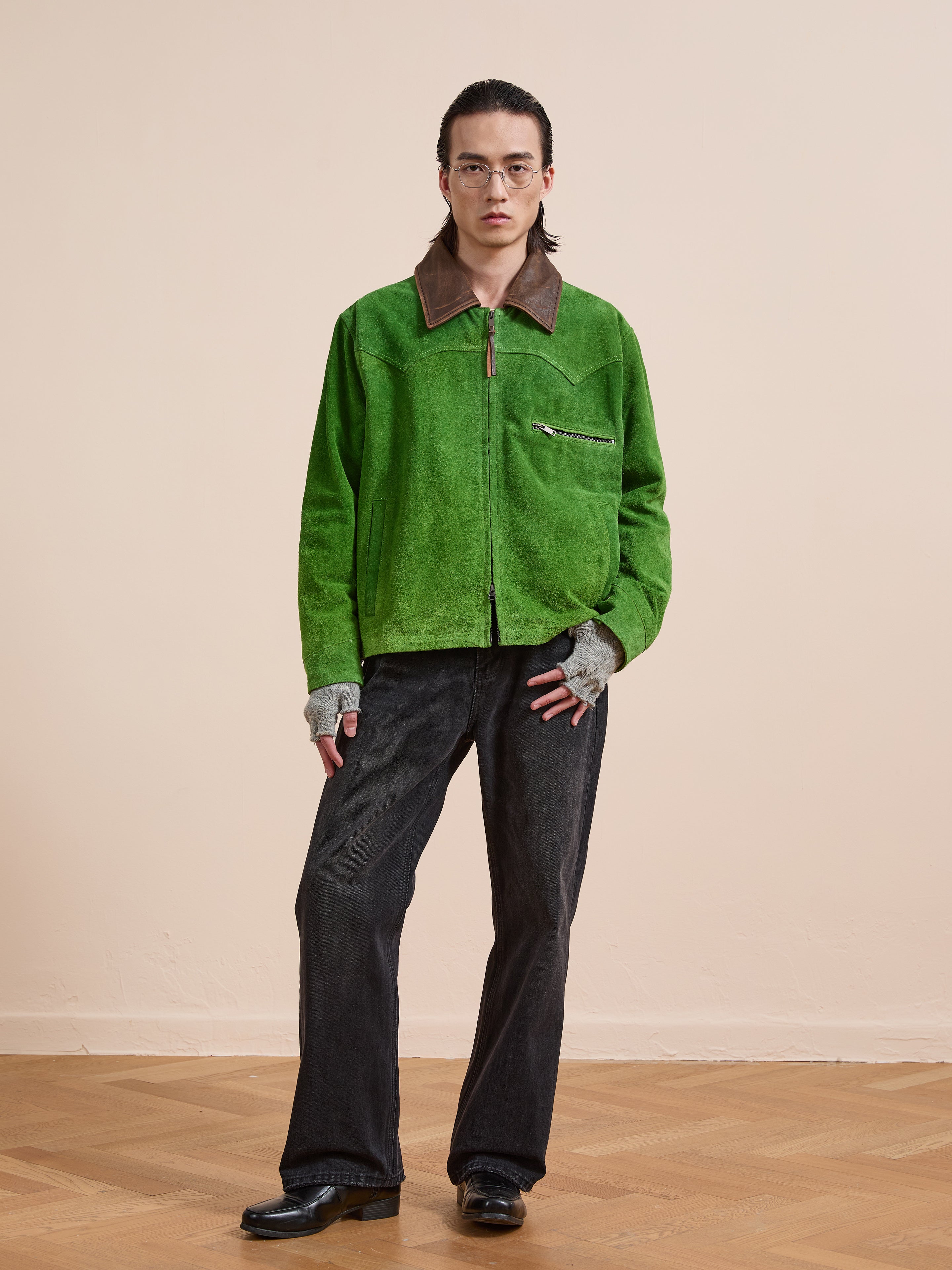 A person in the FOUND Suede Western Barn Jacket, dark jeans, gray gloves, black boots, and glasses stands on a wooden floor against a plain beige background.
