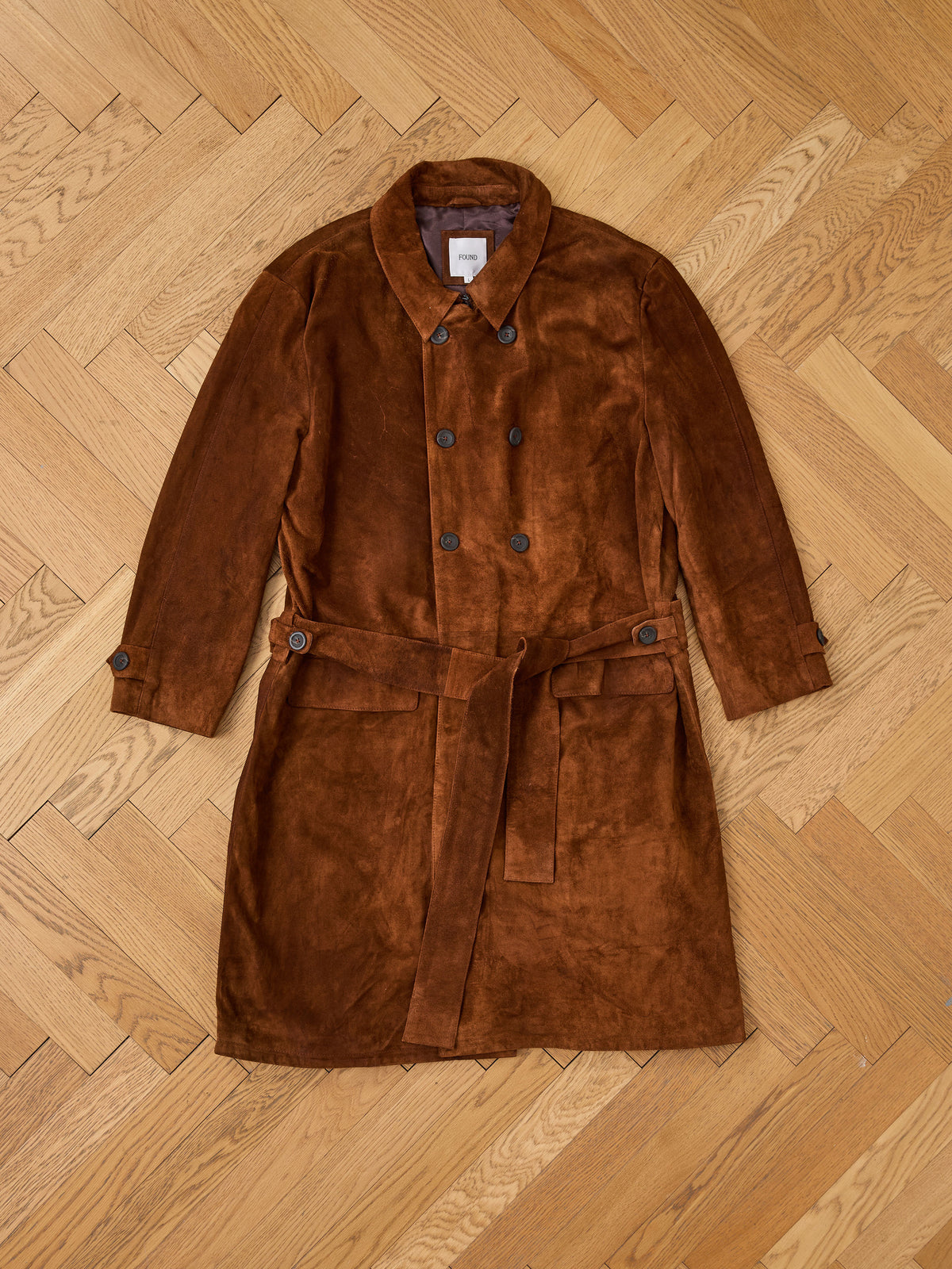 The FOUND Belted Suede Trench Coat, a unisex style in brown suede, is laid flat on a wooden herringbone floor.