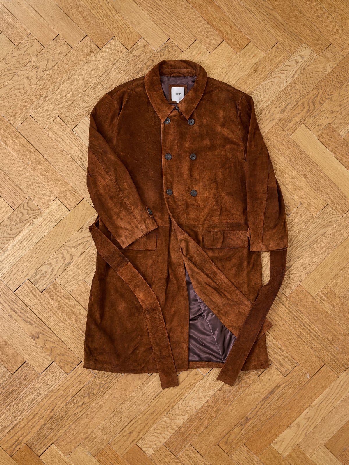 FOUND Belted Suede Trench Coat, unisex and double-breasted, laid flat on a wooden herringbone floor.