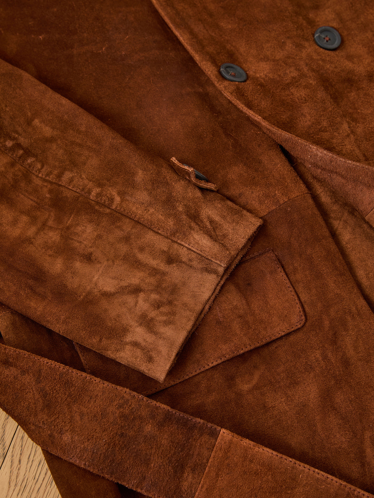 Close-up of the FOUND Belted Suede Trench Coat showing the sleeve, buttons, flap pocket, and belt detail on a wooden surface.