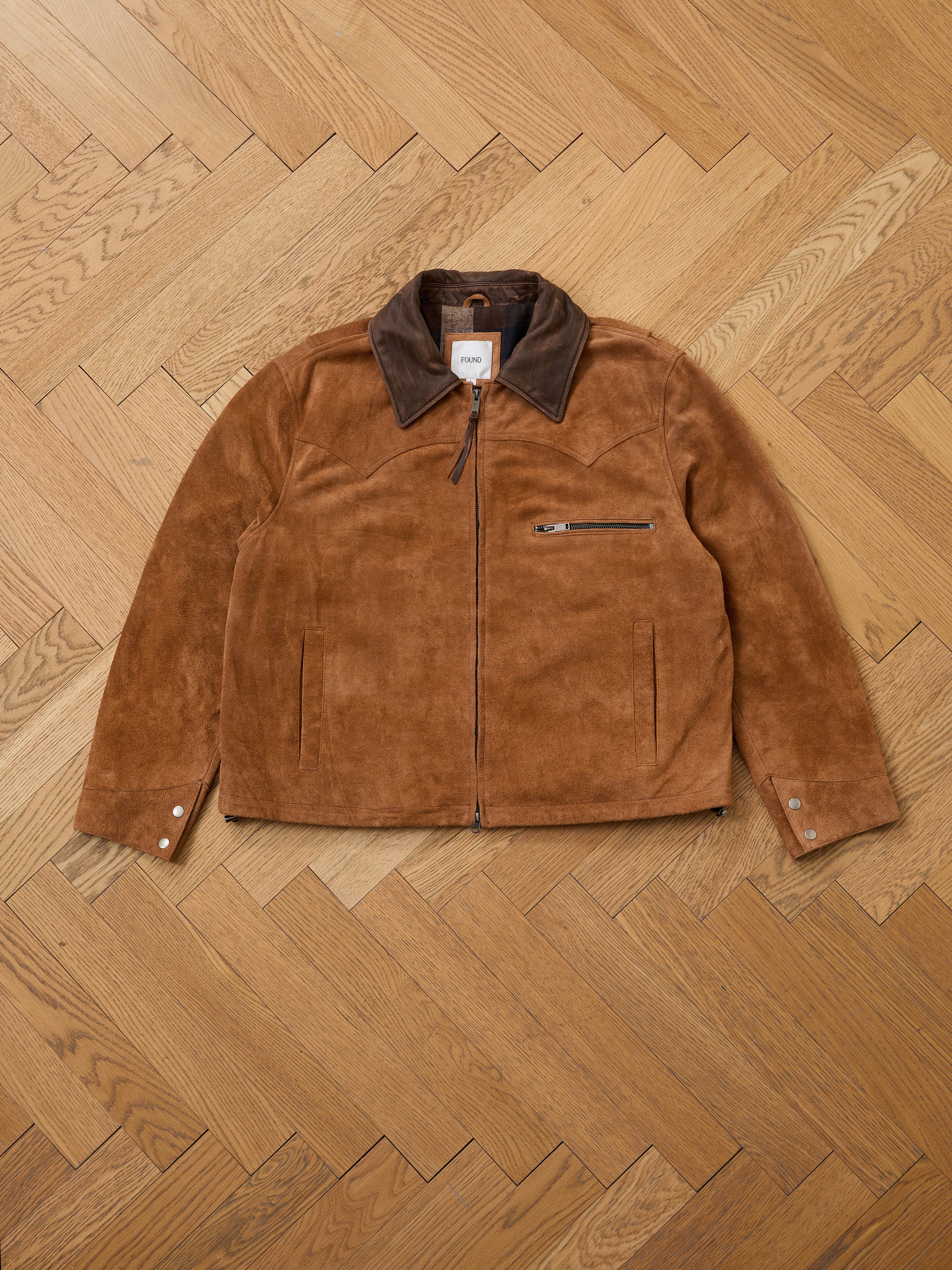 The FOUND Suede Western Barn Jacket features a brown suede body, western yoke stitching, dark brown collar, front zipper, chest zip pocket, two side pockets, and buttoned cuffs, shown laid flat on a herringbone wood floor.