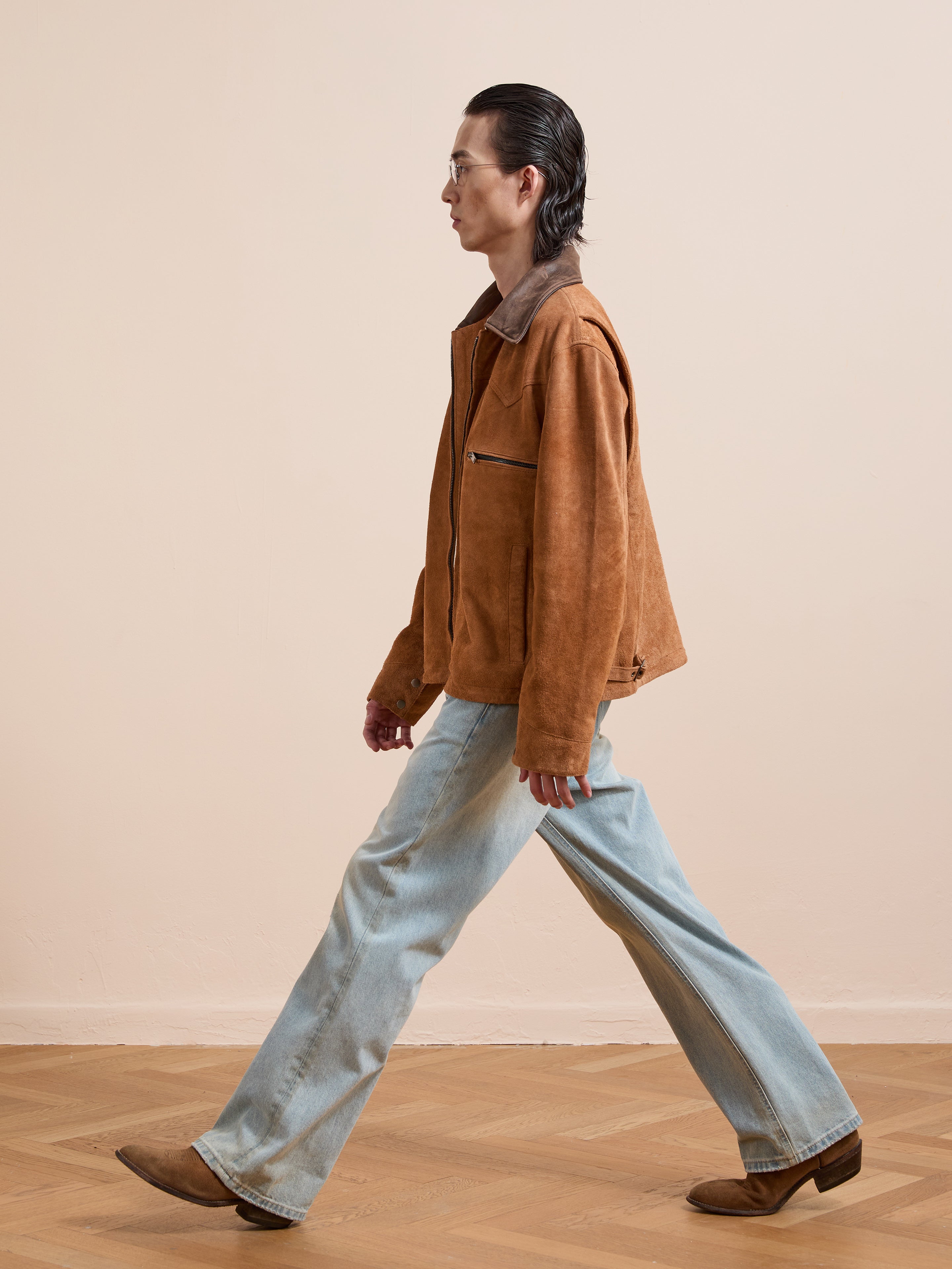 A person with slicked-back hair wears the FOUND Suede Western Barn Jacket, light blue jeans, and brown boots while walking indoors on a wooden floor against a plain beige wall.
