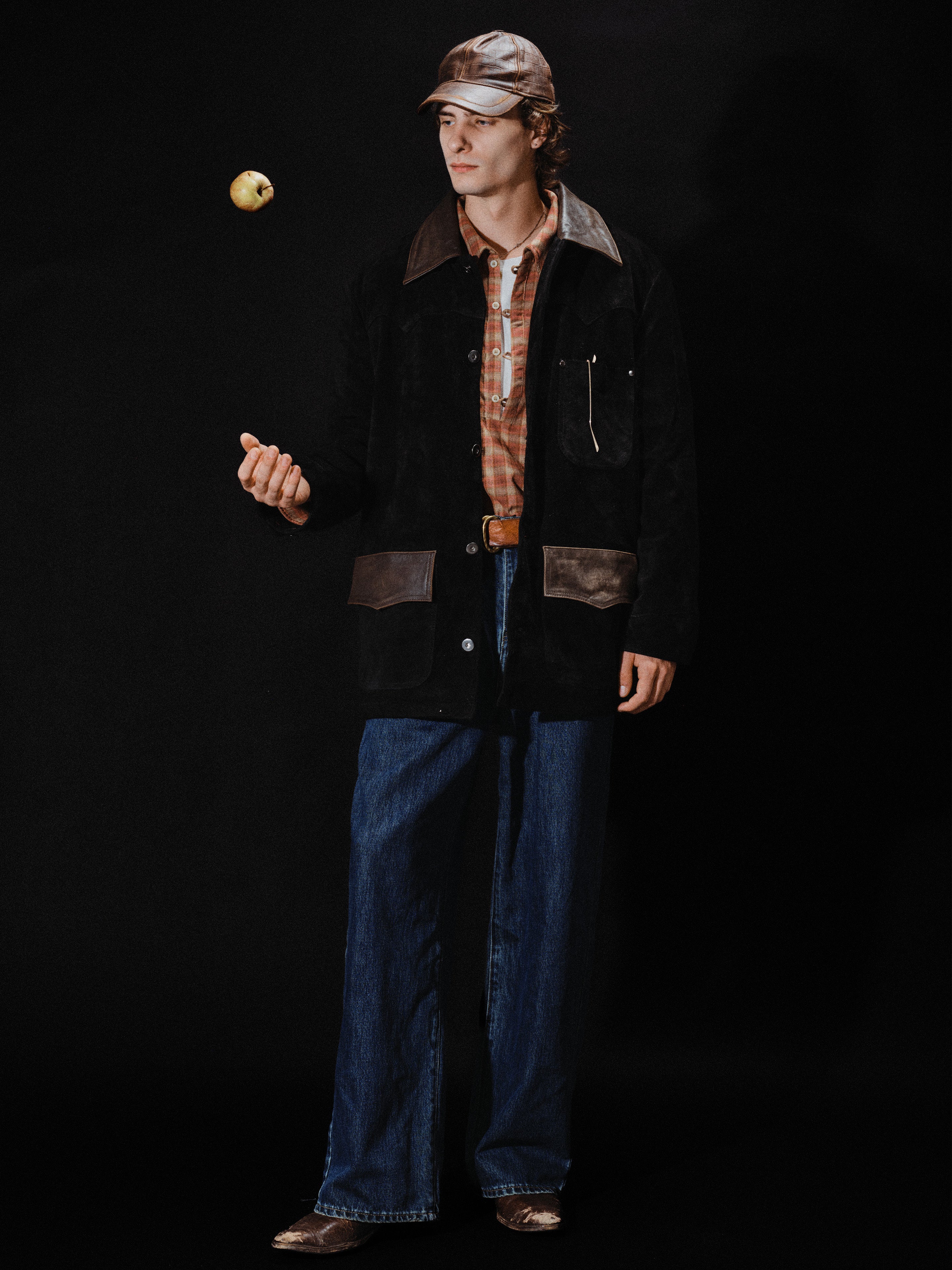 A man wearing the FOUND Suede Contrast Barn Jacket, a plaid shirt, wide-leg jeans, and a cap tosses an apple in the air against a black background.