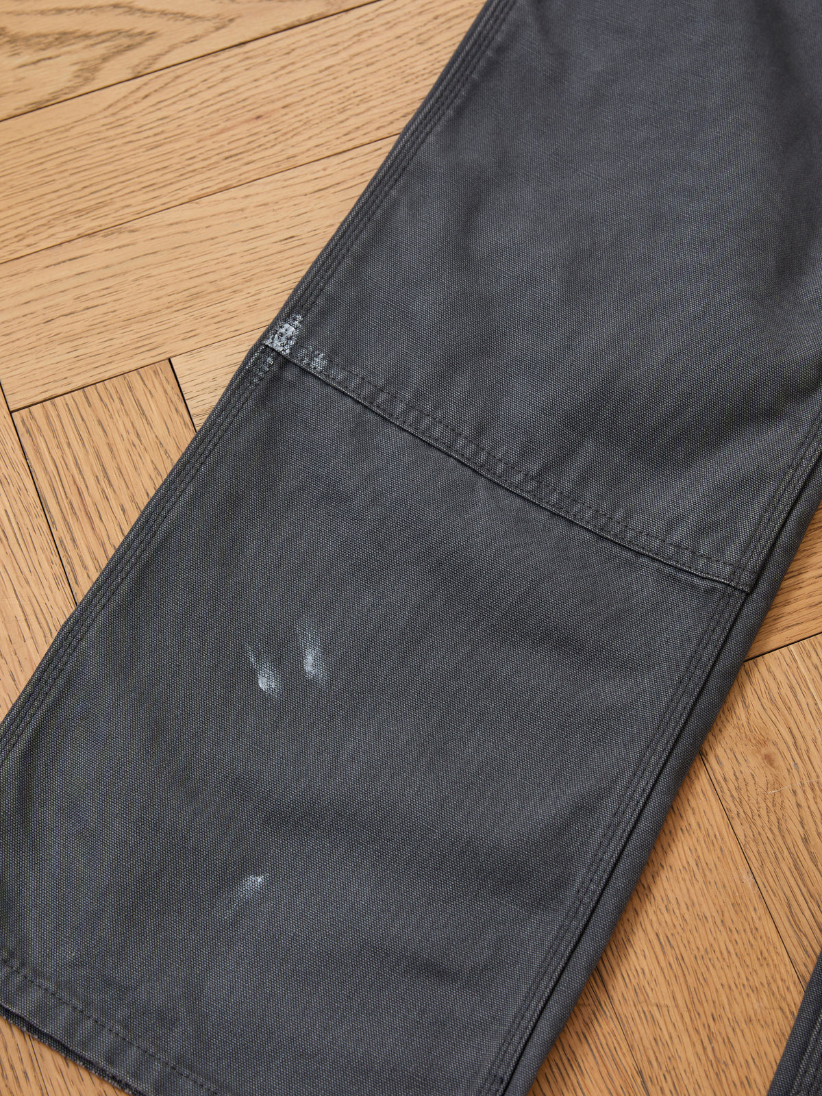Sunbleached Utility Pants by FOUND in dark gray with visible white marks and wear on the knee and thigh, laid flat on a herringbone-patterned wooden floor.