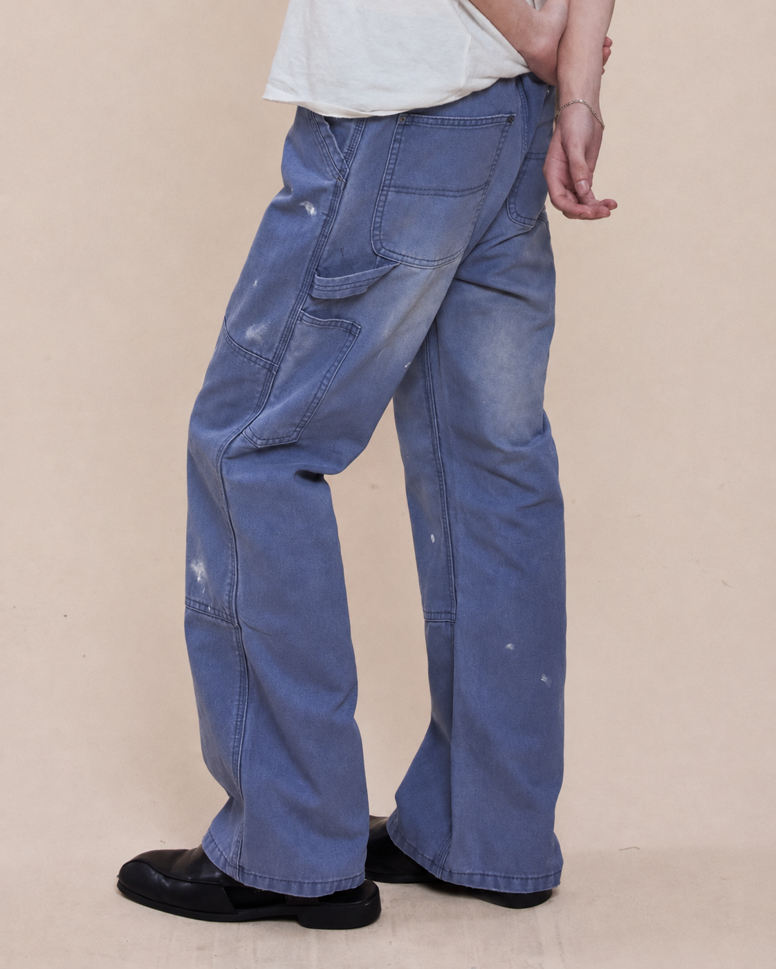 Person in Sunbleached Utility Pants by FOUND—relaxed fit, wide-leg light blue with faded details—paired with black shoes and a white tee against a beige background.
