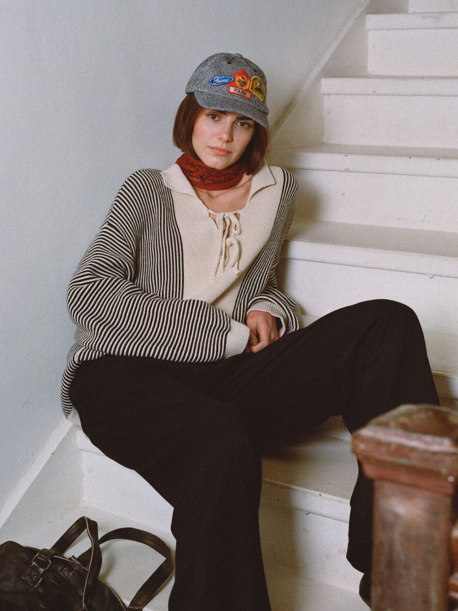 Sitting on the stairs, a person wears a Tabas Tie Knit Collared Sweater by Found, paired with black pants, a cap, and a striped scarf tied around their neck. A bag is placed on the floor beside them.