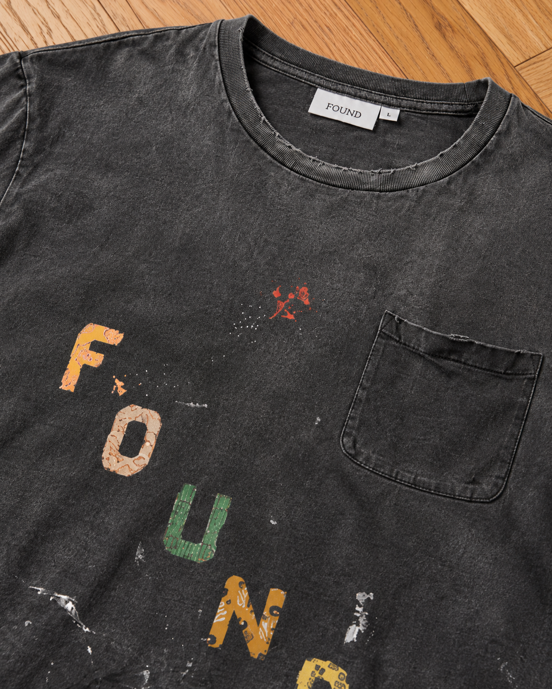 FOUND Tapestry Logo Paint Tee features a chest pocket and faded, multicolored “FOUND” text in a unisex relaxed fit, shown on a wooden surface.