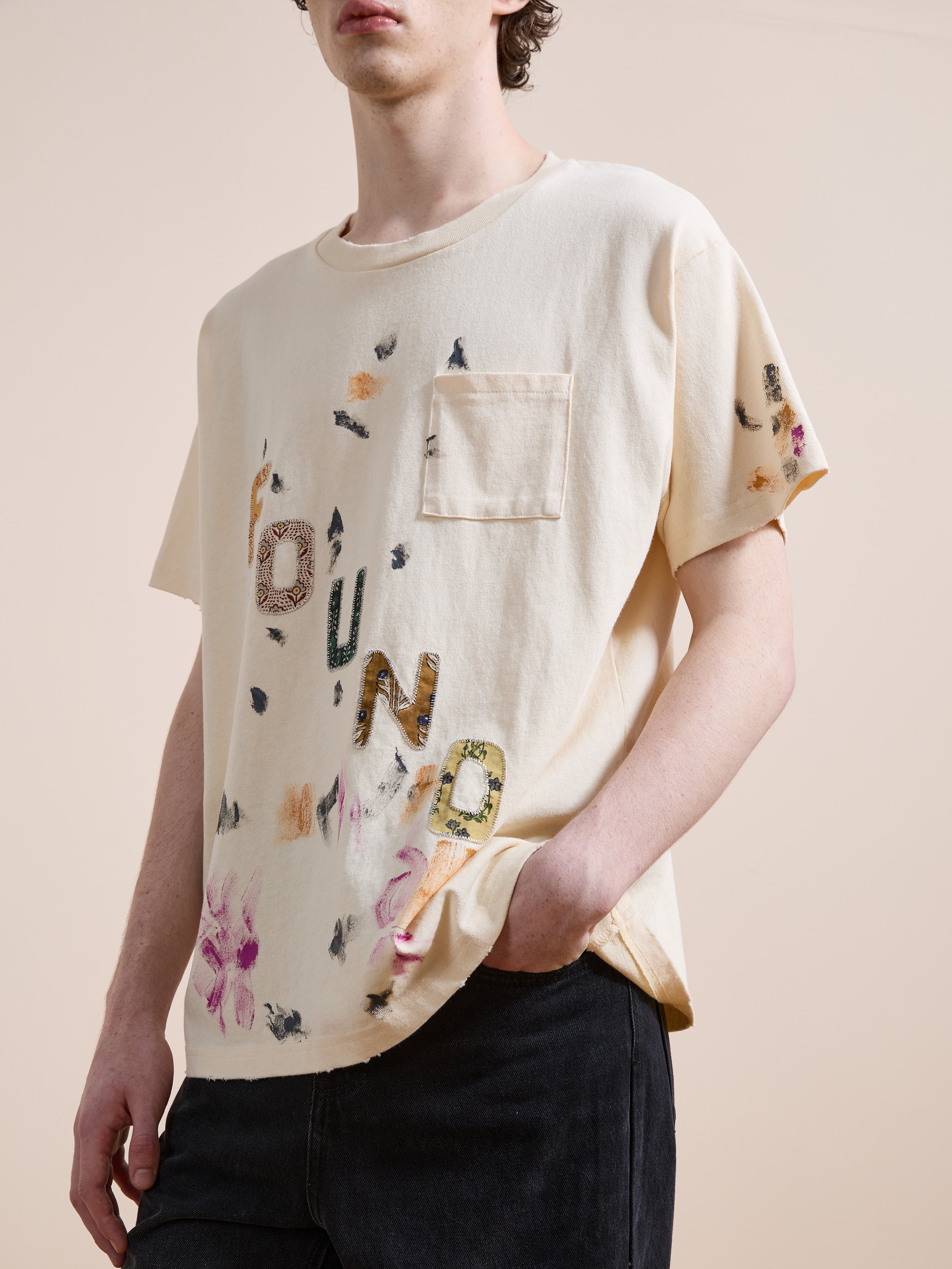 A person wears the FOUND Tapestry Logo Paint Tee—relaxed fit, cream-colored premium cotton with a chest pocket, multicolored floral and logo designs—paired with black pants.