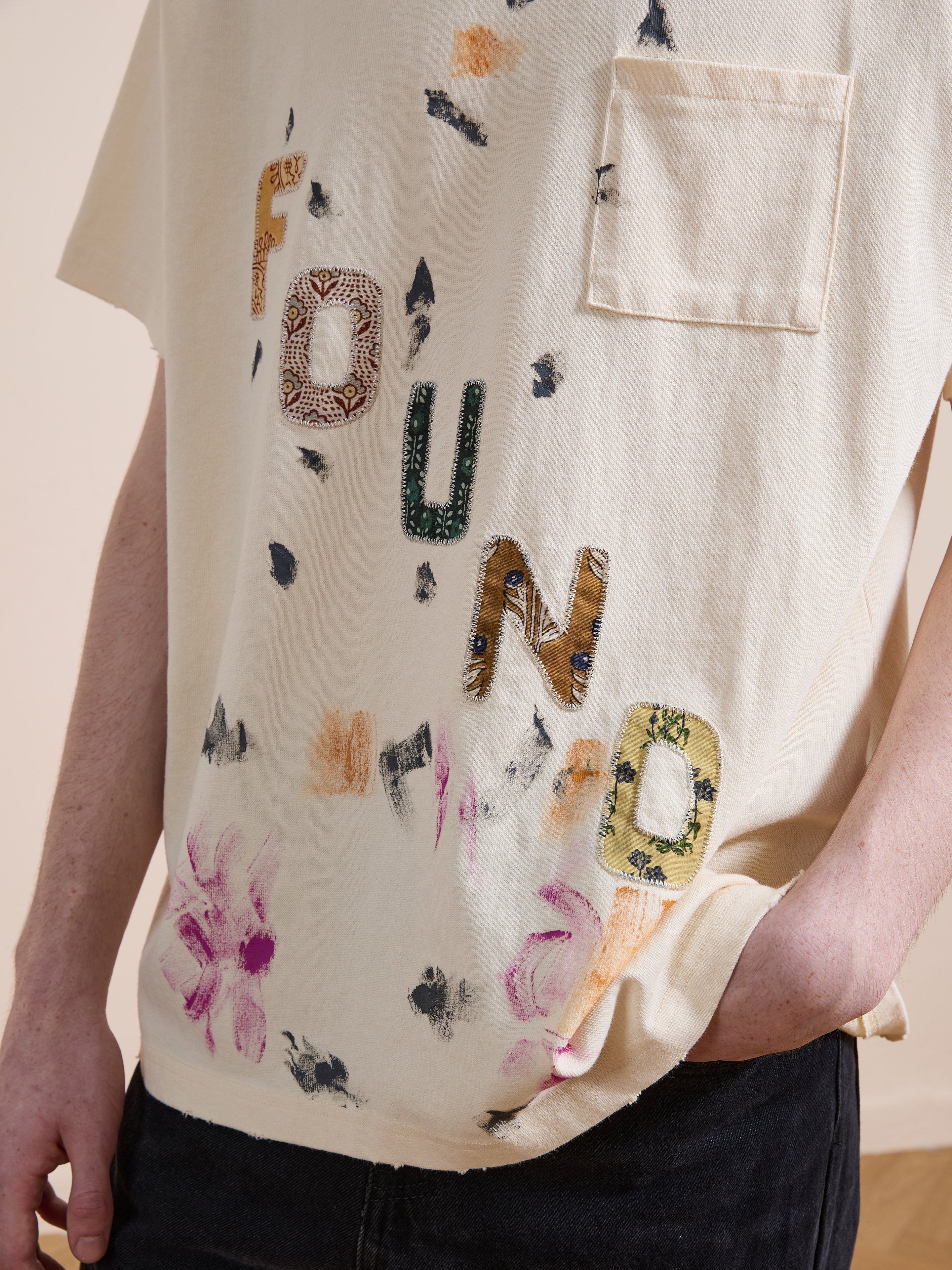 A person wears the FOUND Tapestry Logo Paint Tee, a unisex relaxed fit made from premium cotton, featuring a chest pocket and "FOUND" in colorful textured letters with abstract paint and flower designs.