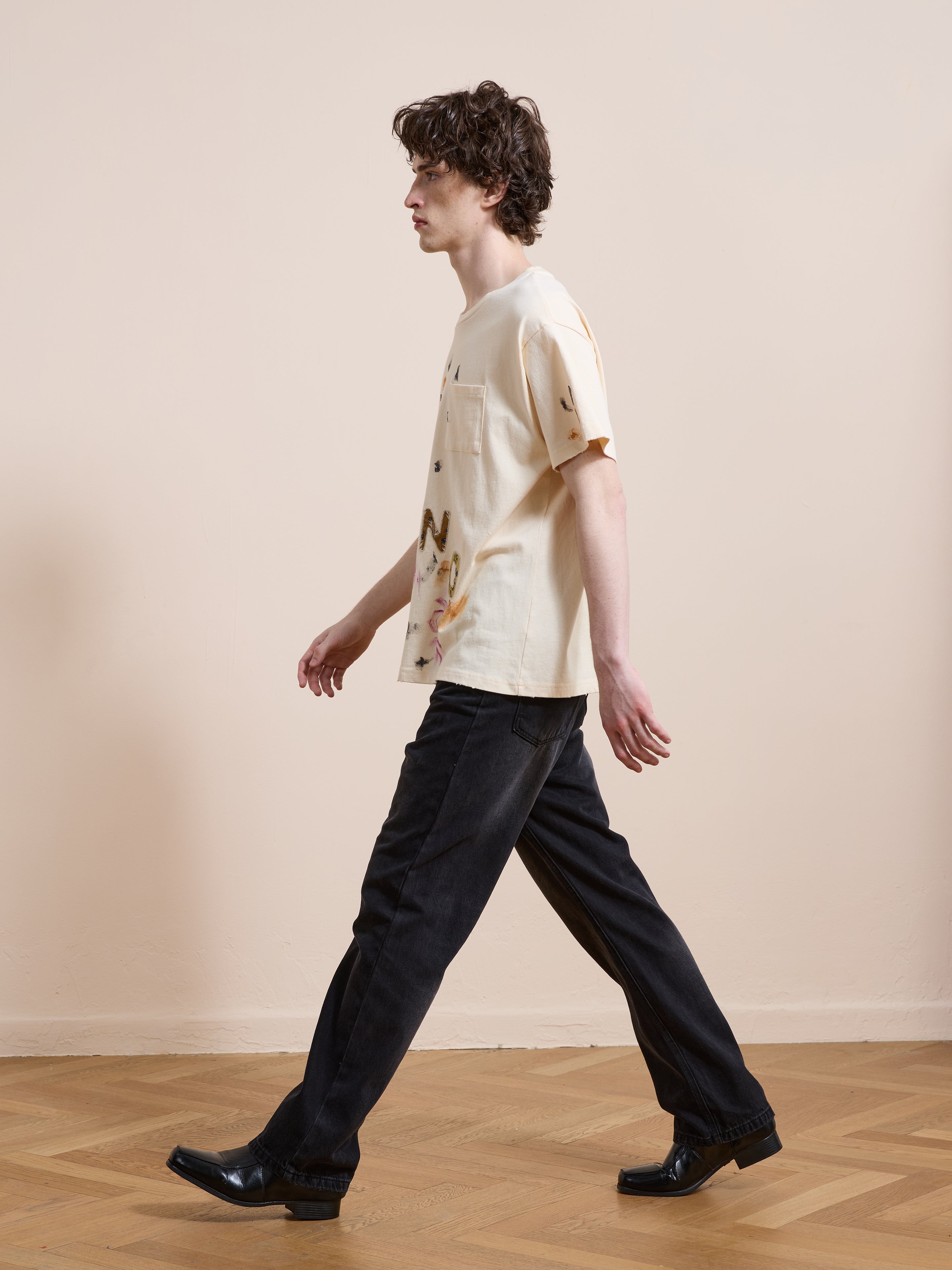 A person with medium-length curly hair walks indoors on a wooden floor, wearing FOUND's Tapestry Logo Paint Tee in a unisex relaxed fit, paired with black jeans and black shoes.