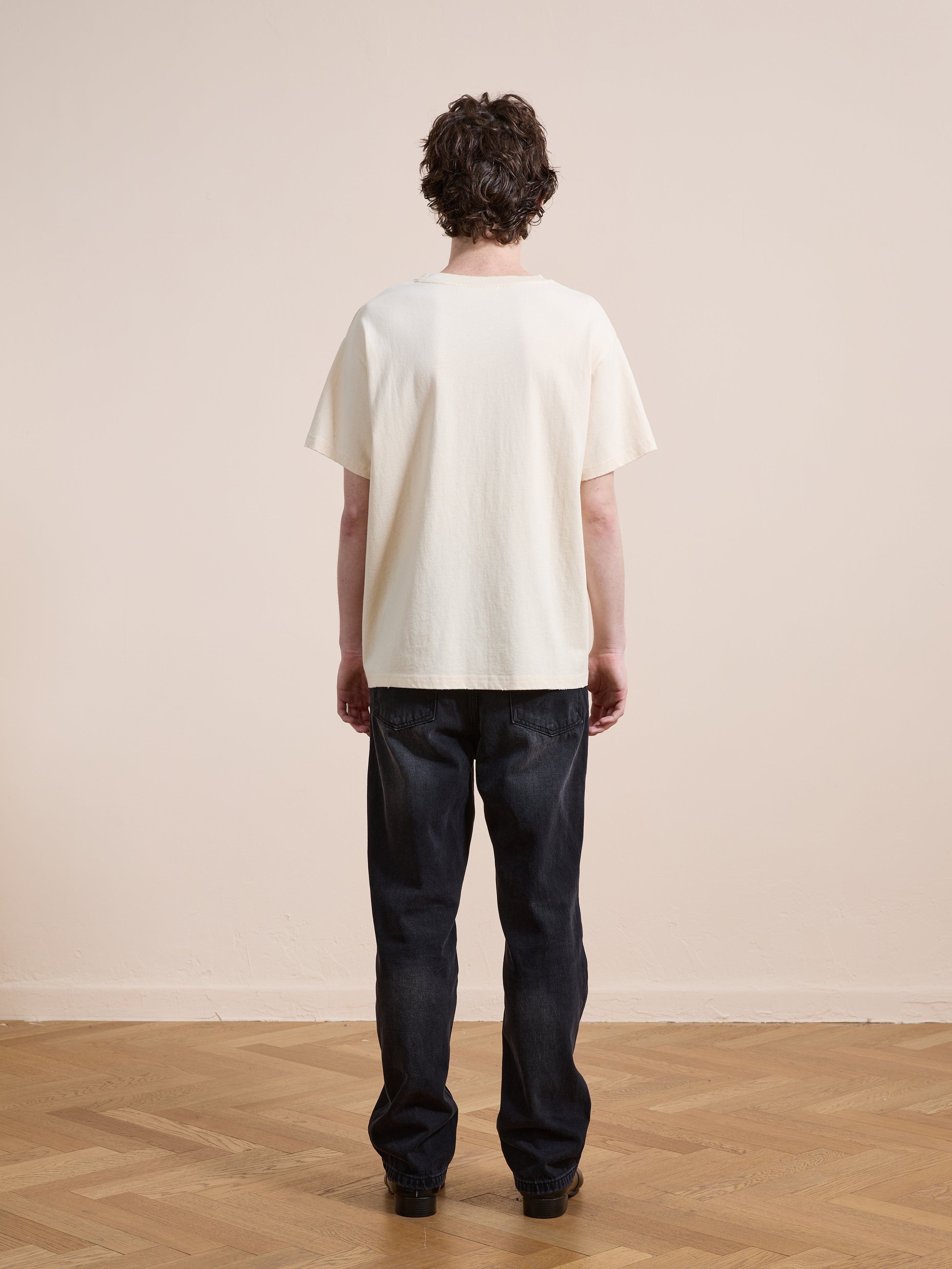 A person with short, curly hair stands indoors facing away, wearing a FOUND Tapestry Logo Paint Tee in white with dark jeans and black shoes on a wooden floor against a neutral background.