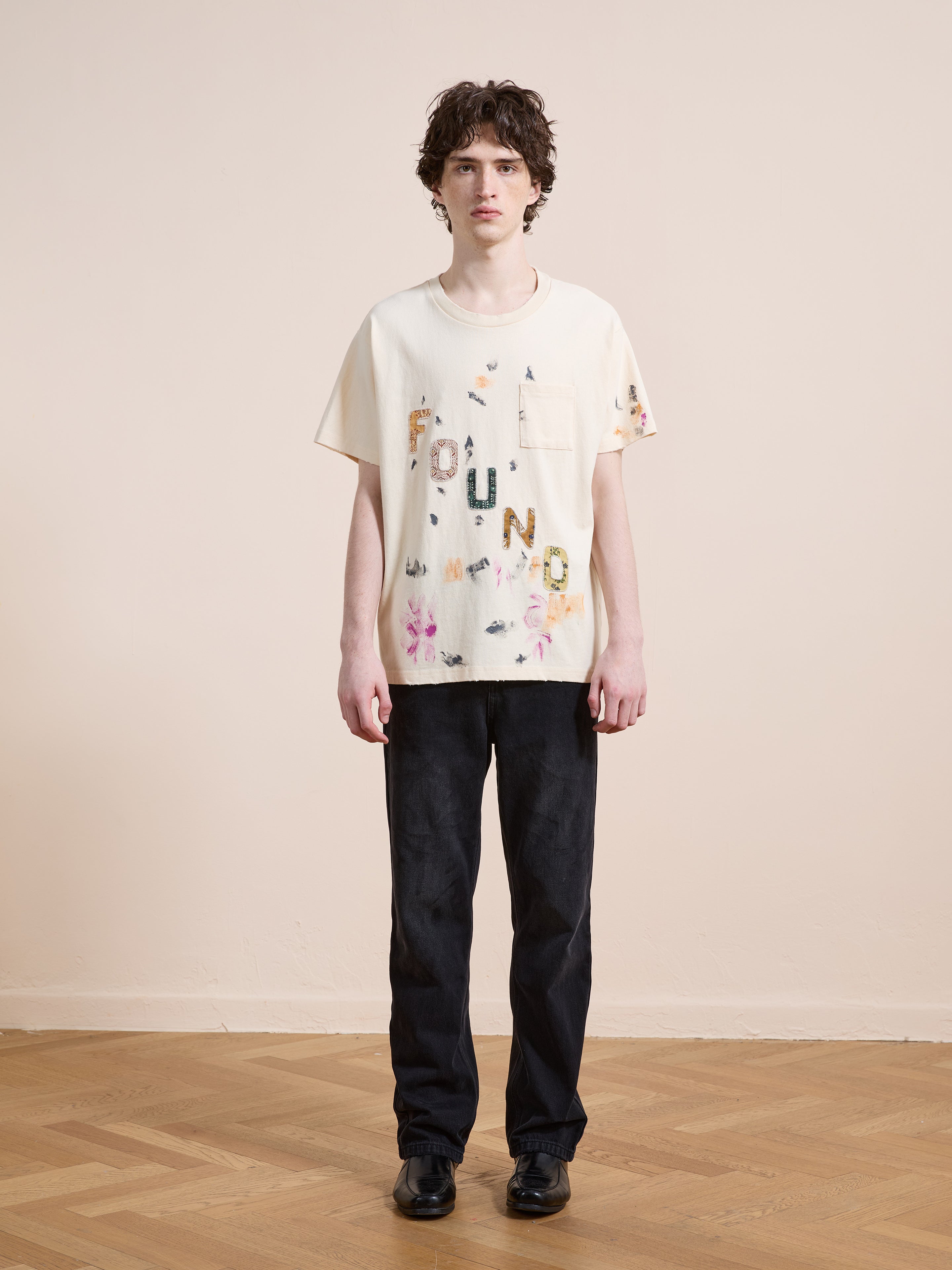 A person stands indoors on a wooden floor, facing the camera against a plain light wall, wearing the unisex relaxed fit FOUND Tapestry Logo Paint Tee in cream with floral designs, paired with dark pants and black shoes.