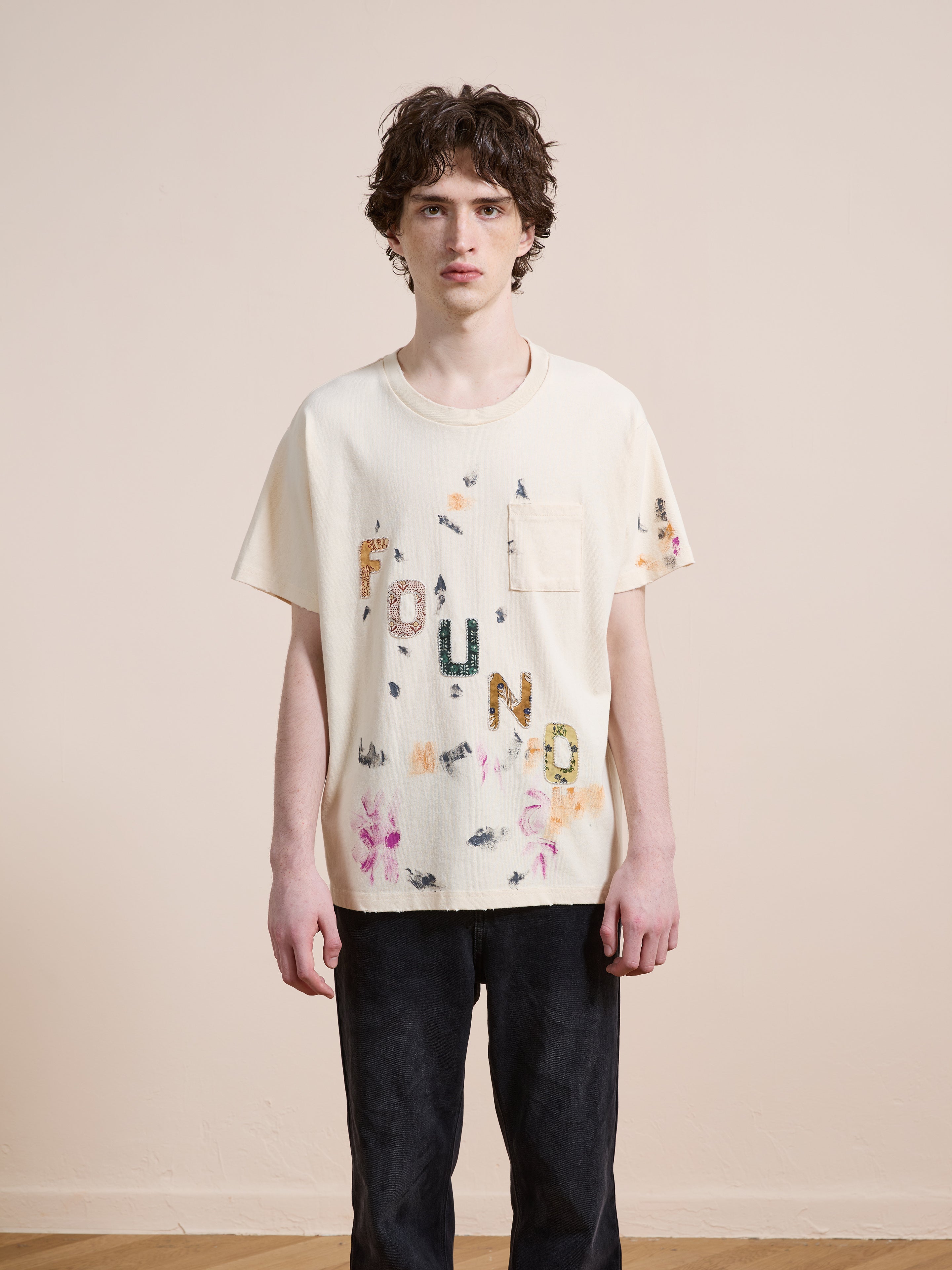 A young person wears the FOUND Tapestry Logo Paint Tee, a cream-colored premium cotton shirt with a small pocket and colorful "FOUND" patches, styled with dark pants for a relaxed unisex look.