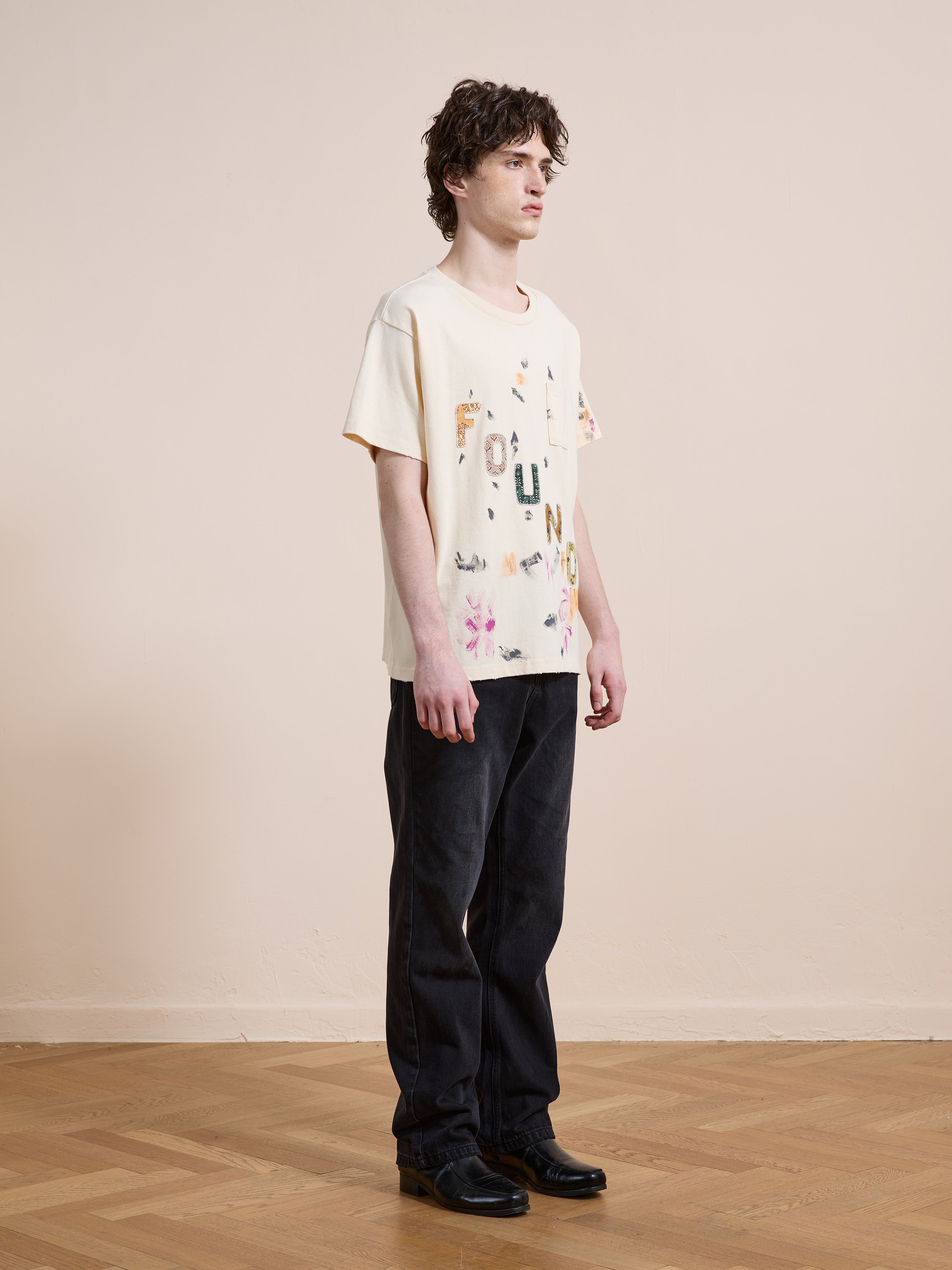 A person stands indoors on a wooden floor, facing slightly right against a beige wall, wearing FOUND's cream Tapestry Logo Paint Tee in premium cotton with colorful letters and black pants.