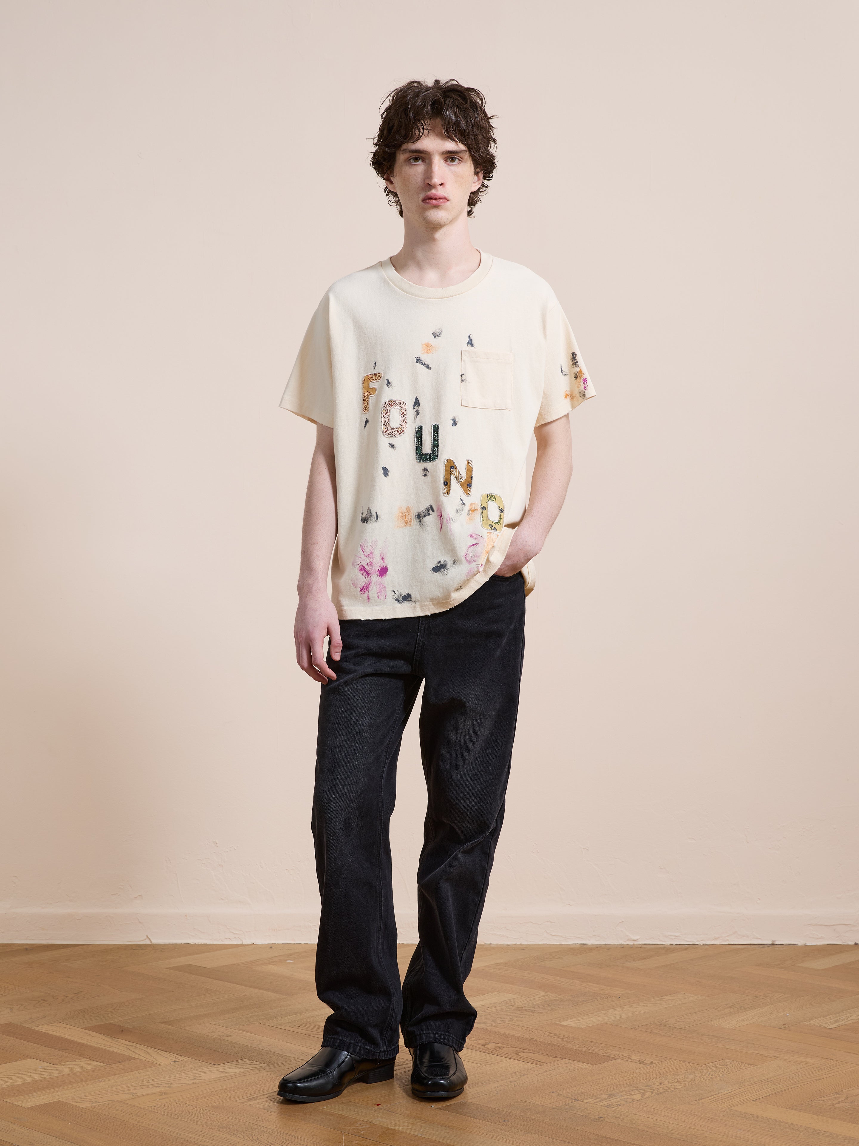 A person stands indoors on a wooden floor wearing the FOUND Tapestry Logo Paint Tee in cream premium cotton, paired with black pants and shoes. The unisex tee features a relaxed fit and bold 'FOUND' branding, with one hand casually in their pocket.