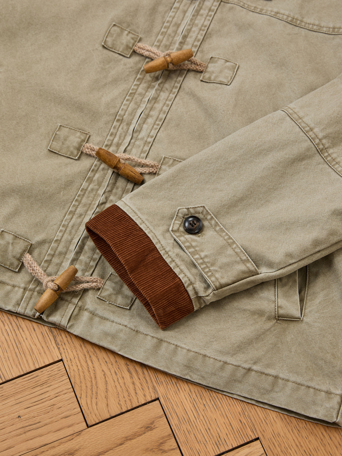 Close-up of the Toggle Work Jacket by FOUND in beige with brown corduroy cuff, button detail, wooden toggles, and vintage-inspired visible stitching on parquet.