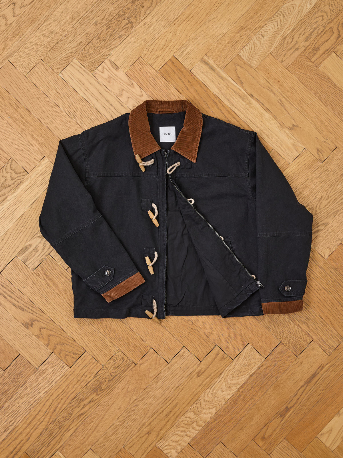 The Found Toggle Work Jacket features a black vintage design with a brown corduroy collar and cuffs, fireman-style toggle closure, and an inside tag, displayed on a herringbone wood floor.