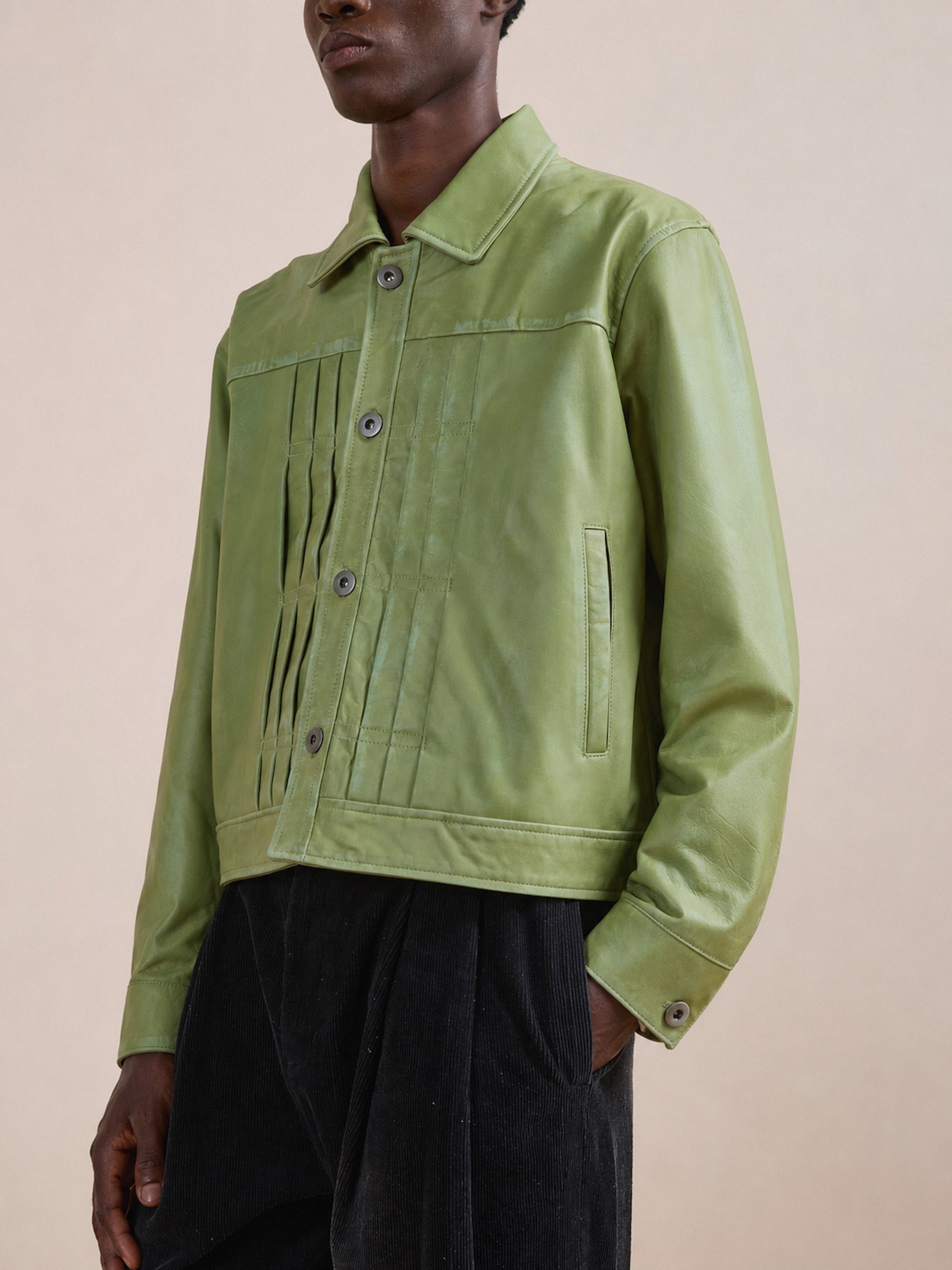 A person is wearing the FOUND Trucker Leather Jacket in light green, featuring vertical pleats on the front and a unisex relaxed fit, paired with black corduroy pants and standing against a neutral background.