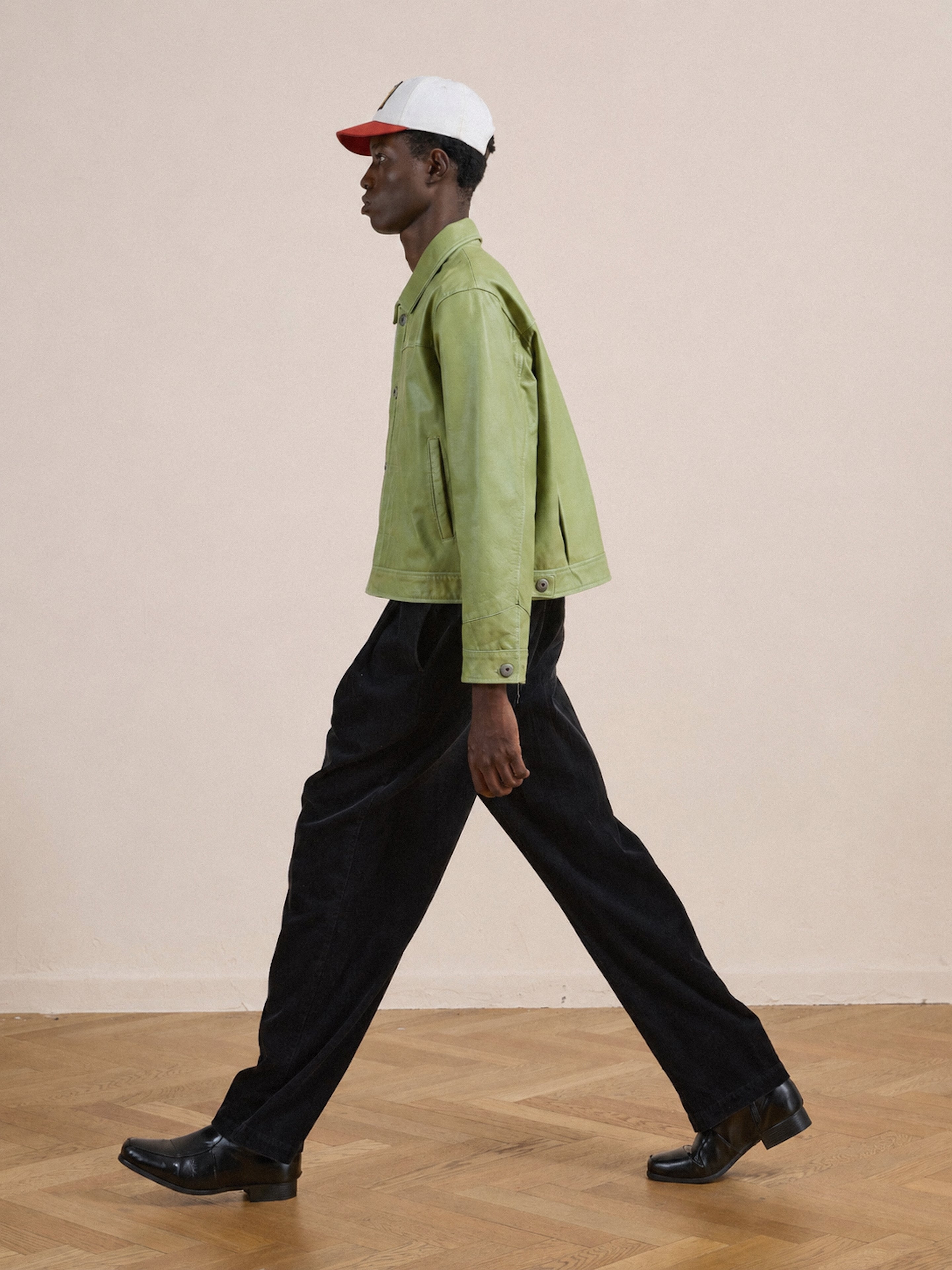 Wearing the FOUND Trucker Leather Jacket in light green with a unisex relaxed fit, paired with black pants, black shoes, and a white and red cap, a person walks across a wooden floor against a plain beige background.