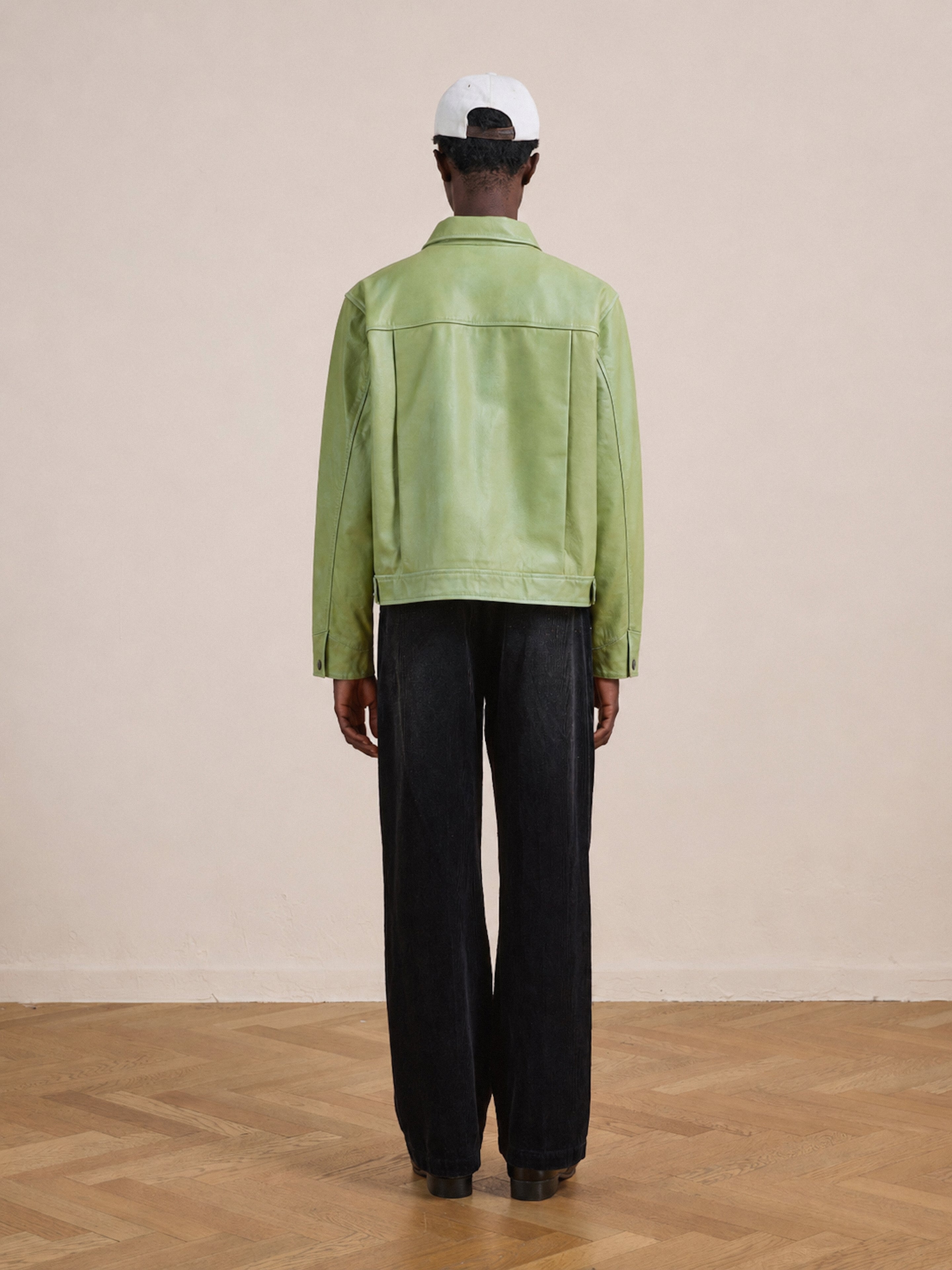 A person stands indoors facing away, wearing the FOUND Trucker Leather Jacket in light green with a unisex relaxed fit, paired with black pants, black shoes, and a white cap. The room features a light-colored wall and a wooden floor.