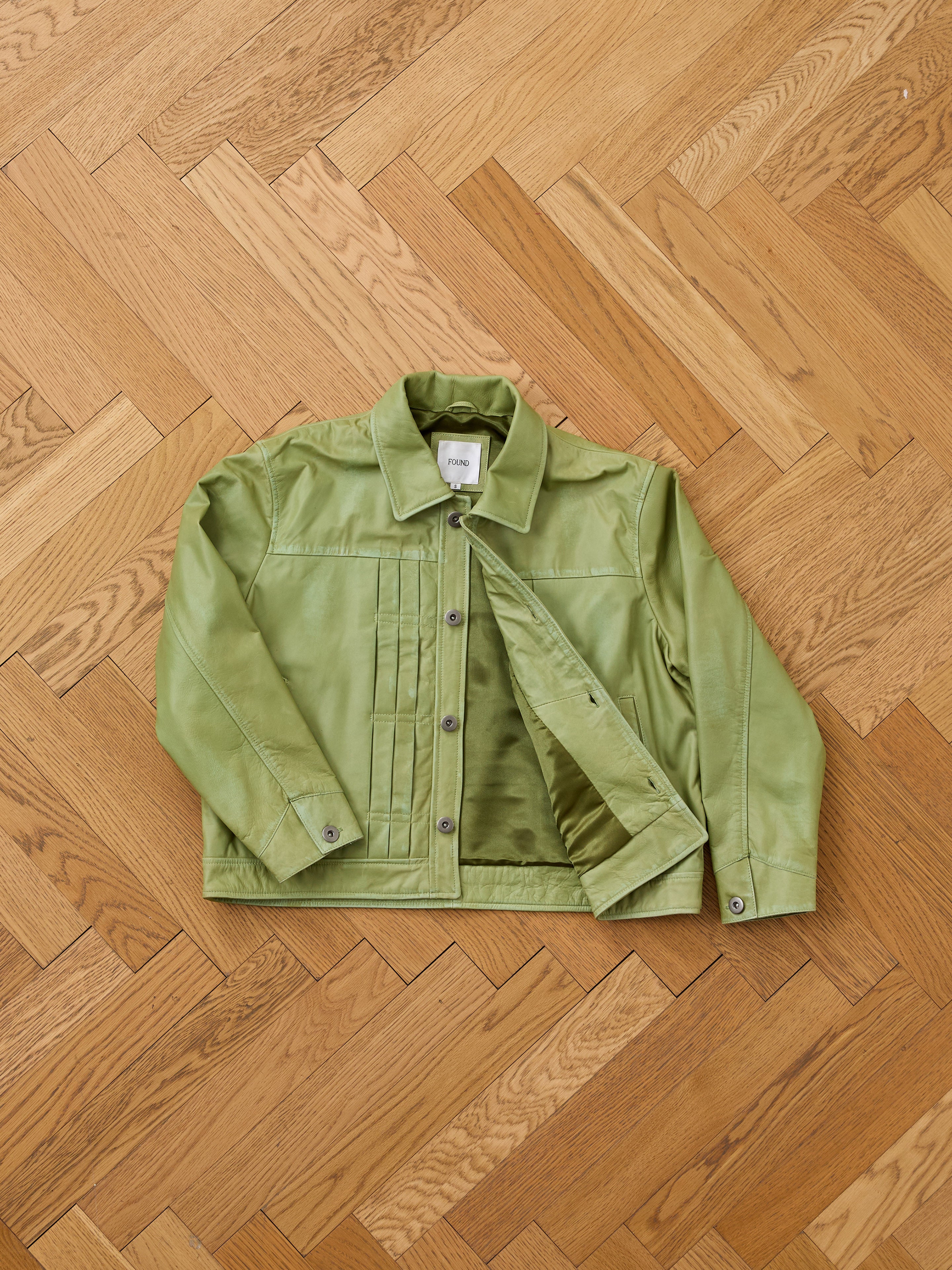 The FOUND Trucker Leather Jacket in washed green leather, with button closure and pleated detailing, is displayed unisex and laid flat on a light wooden herringbone floor.